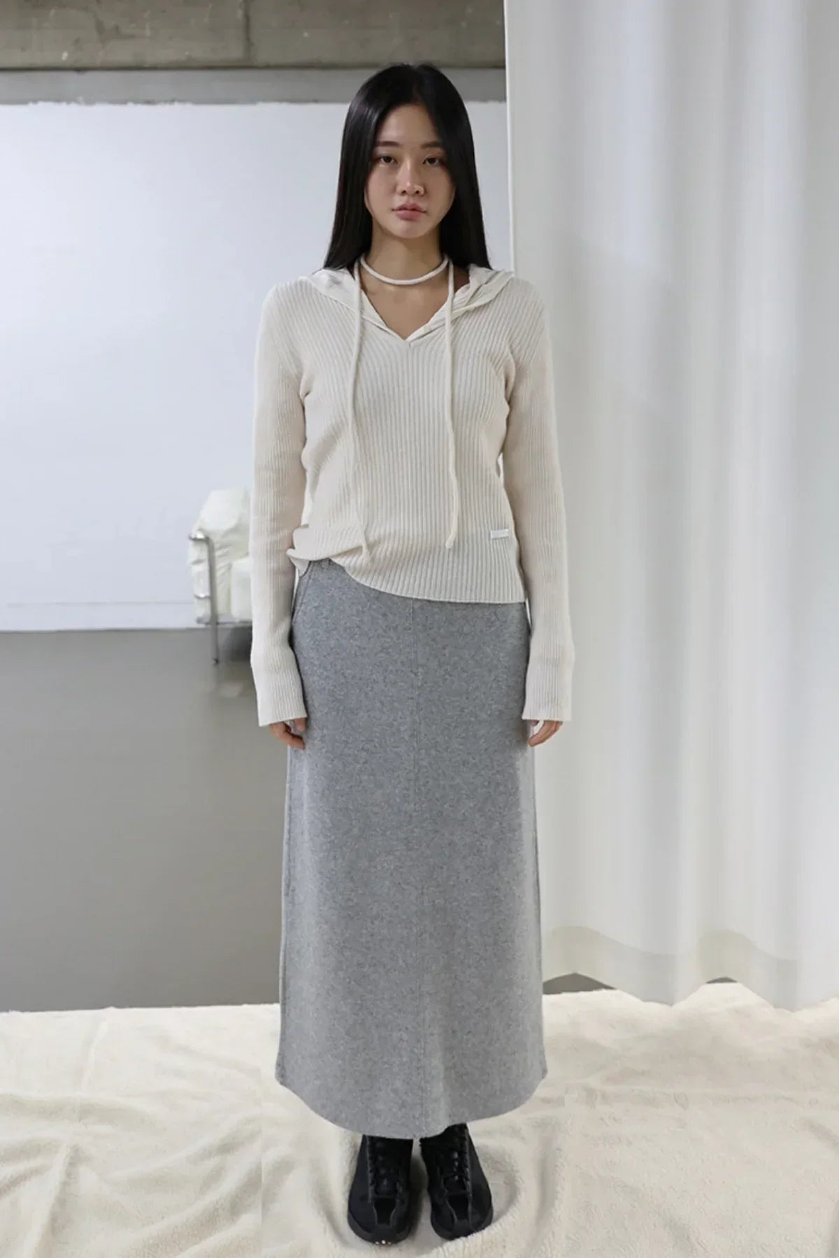 Woman wearing ivory ribbed hoodie and grey long skirt with black lace-up shoes in minimal indoor setting