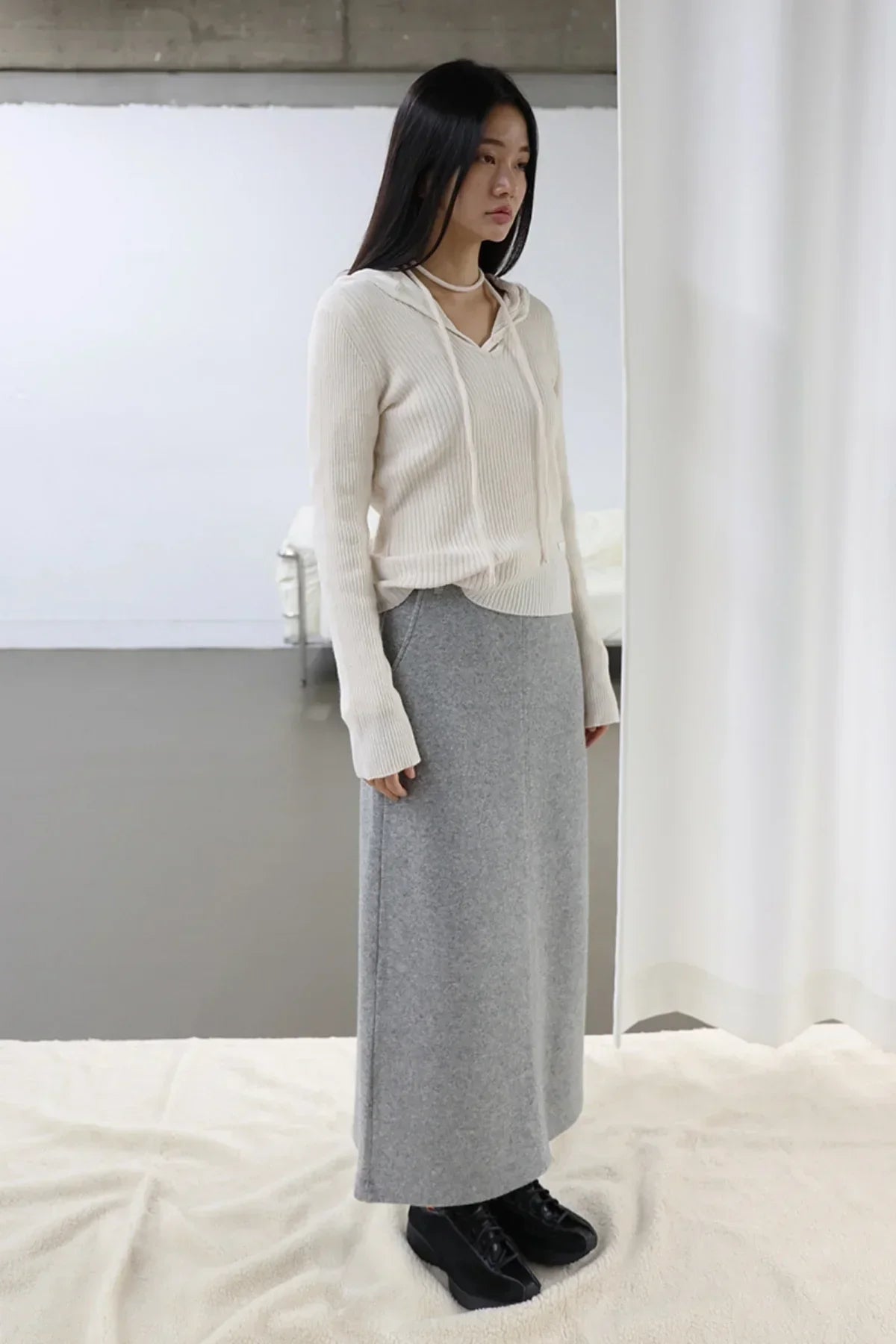 Woman wearing ivory ribbed cashmere hoodie sweater and long grey skirt with black shoes in minimal indoor setting