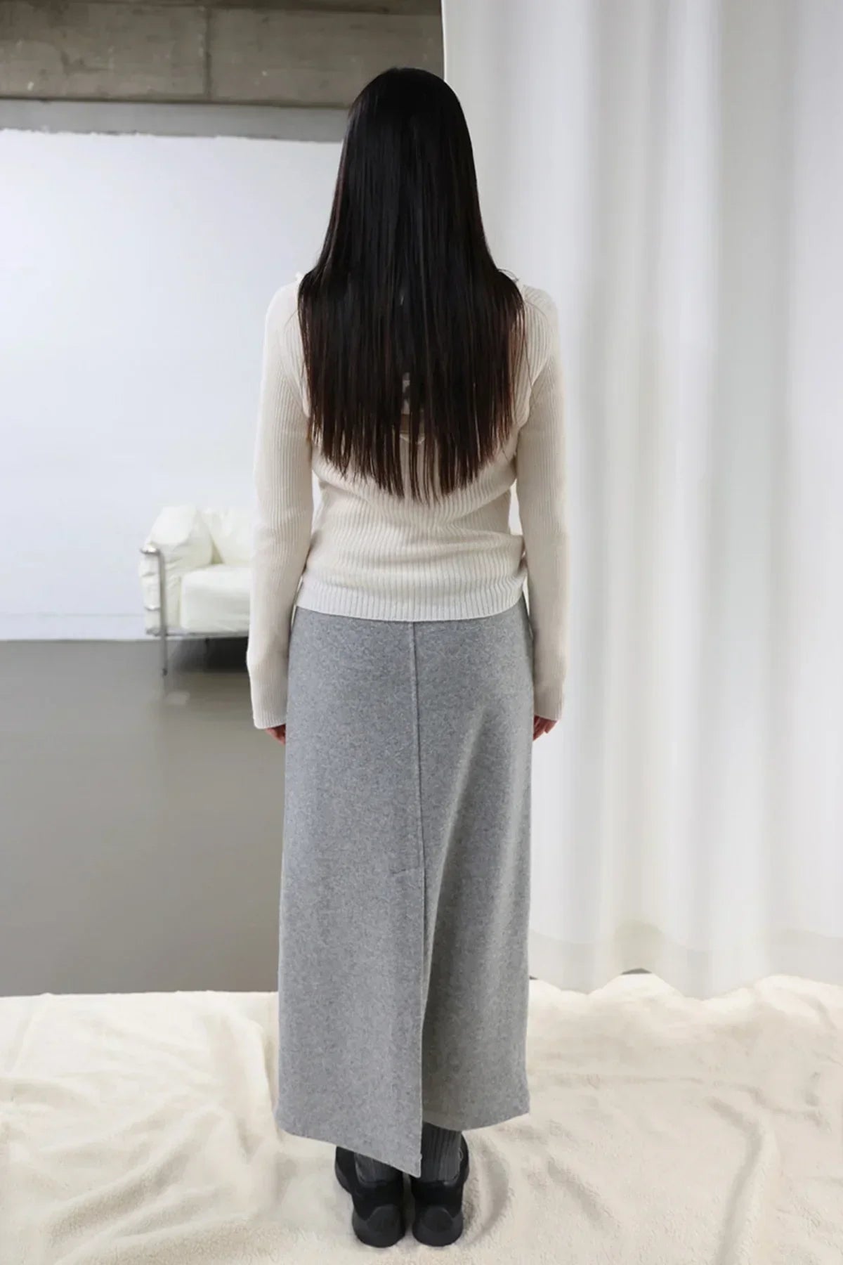 Rear view of woman with long hair wearing an ivory ribbed knit sweater and gray wool midi skirt indoors