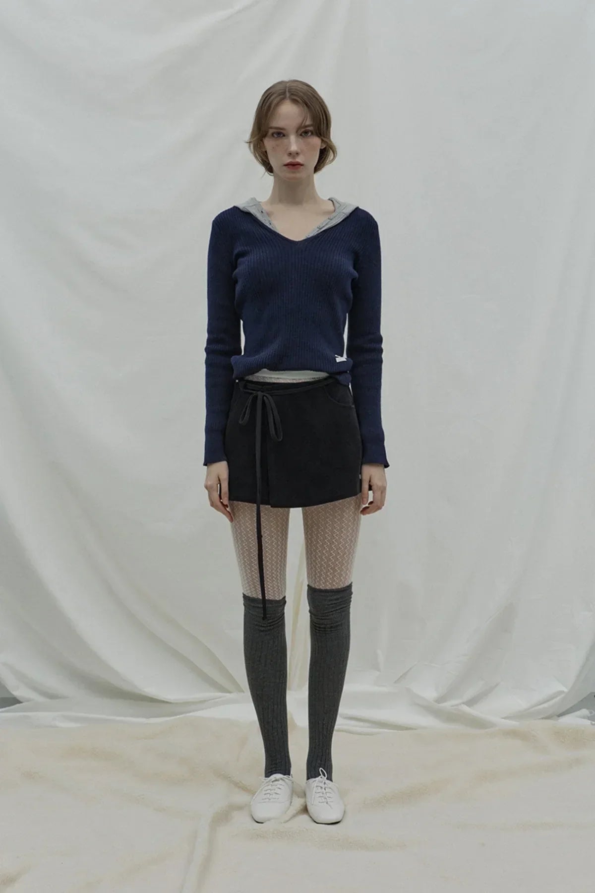 Woman wearing navy v-neck cashmere knit, black skirt, patterned tights, gray knee-high socks, and white shoes