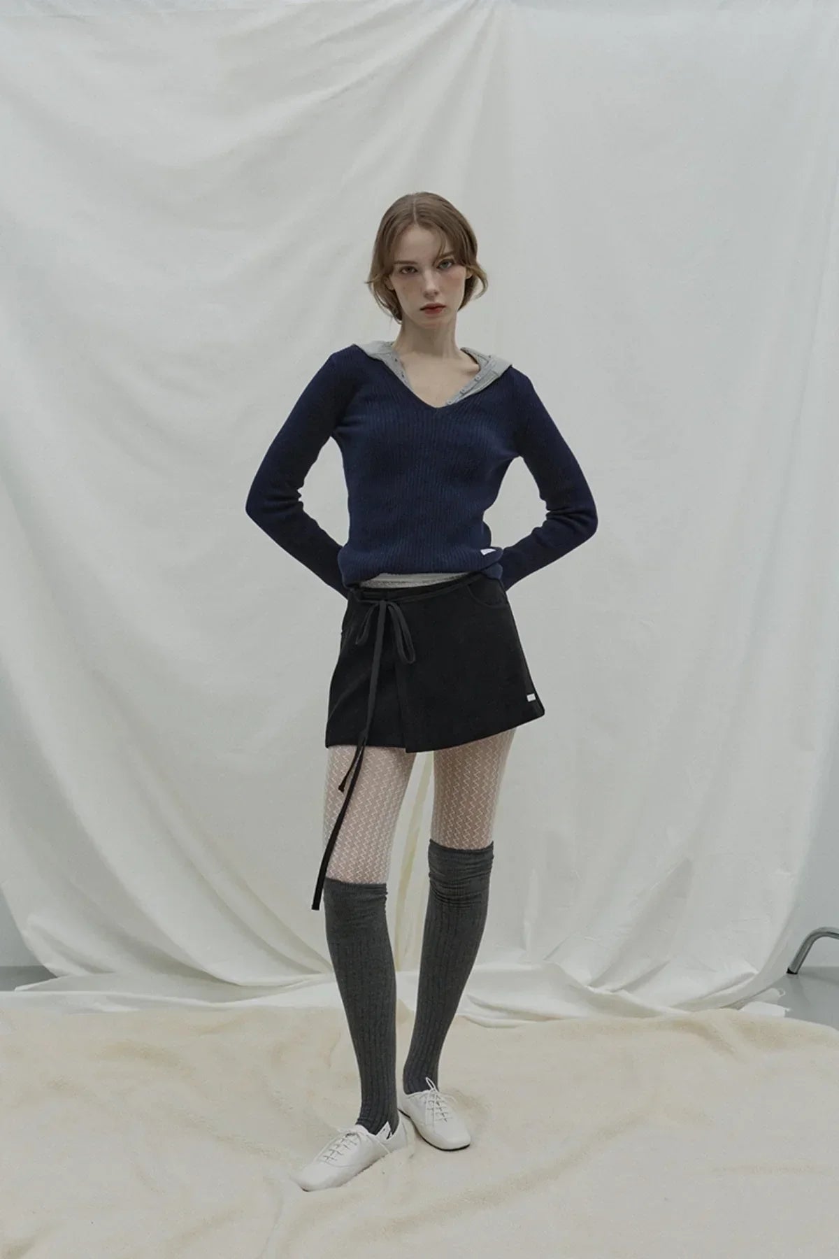 Model wearing navy v-neck cashmere sweater over gray hoodie, black skirt, white patterned tights, gray knee-high socks, and white shoes