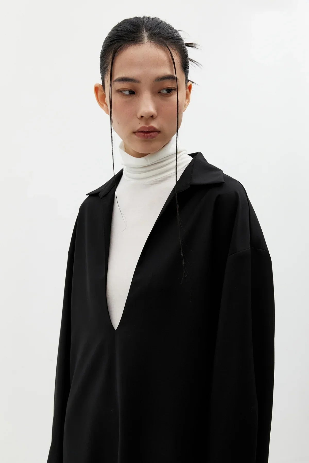 Young woman wearing black oversized V-neck dress layered over white turtleneck against white background