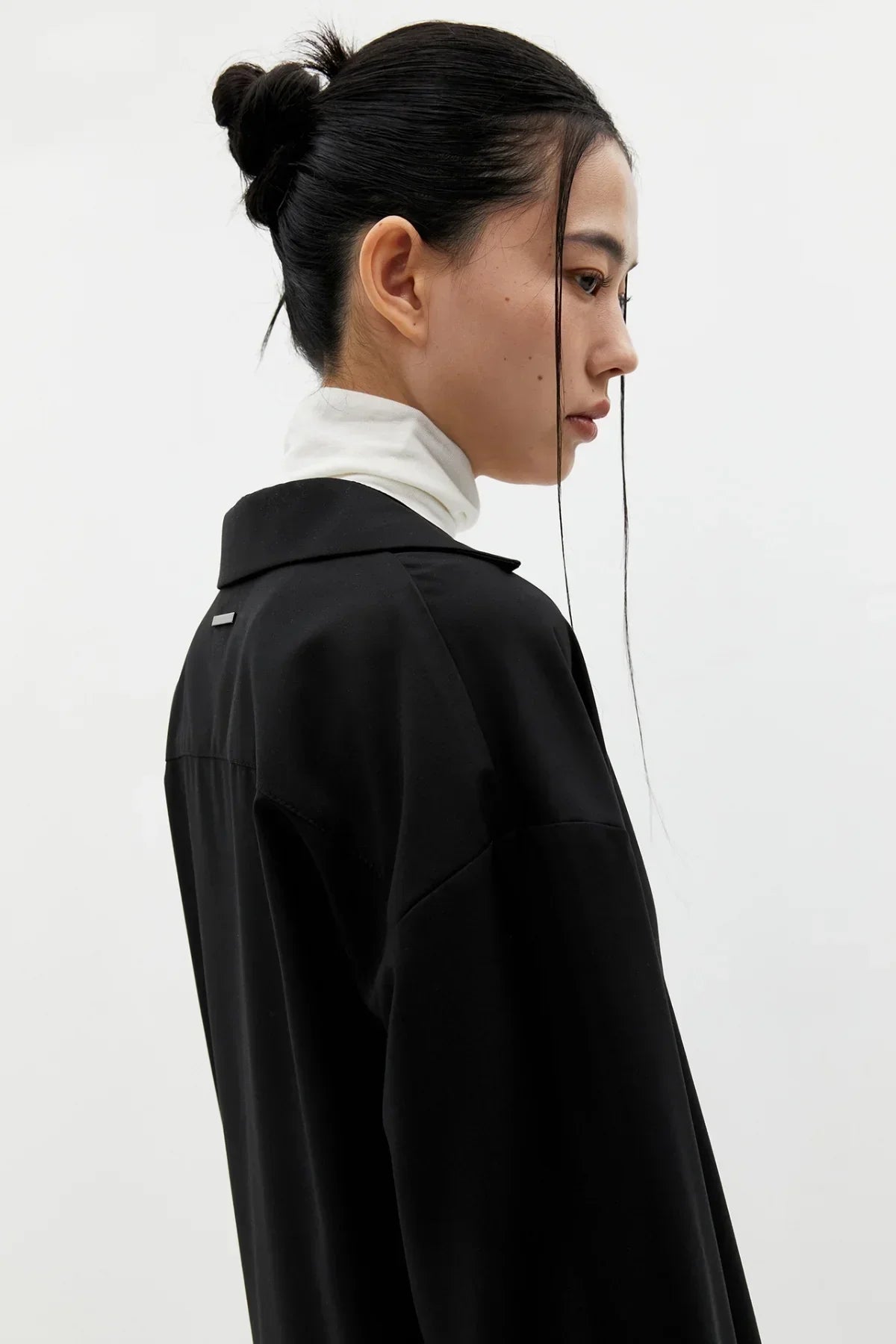 Side profile of woman with black hair bun wearing black oversized jacket and white turtleneck