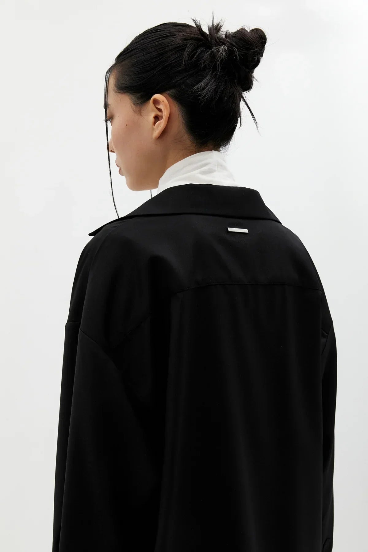 Side profile of a woman with black hair in a bun wearing a black jacket over a white top against a plain light background