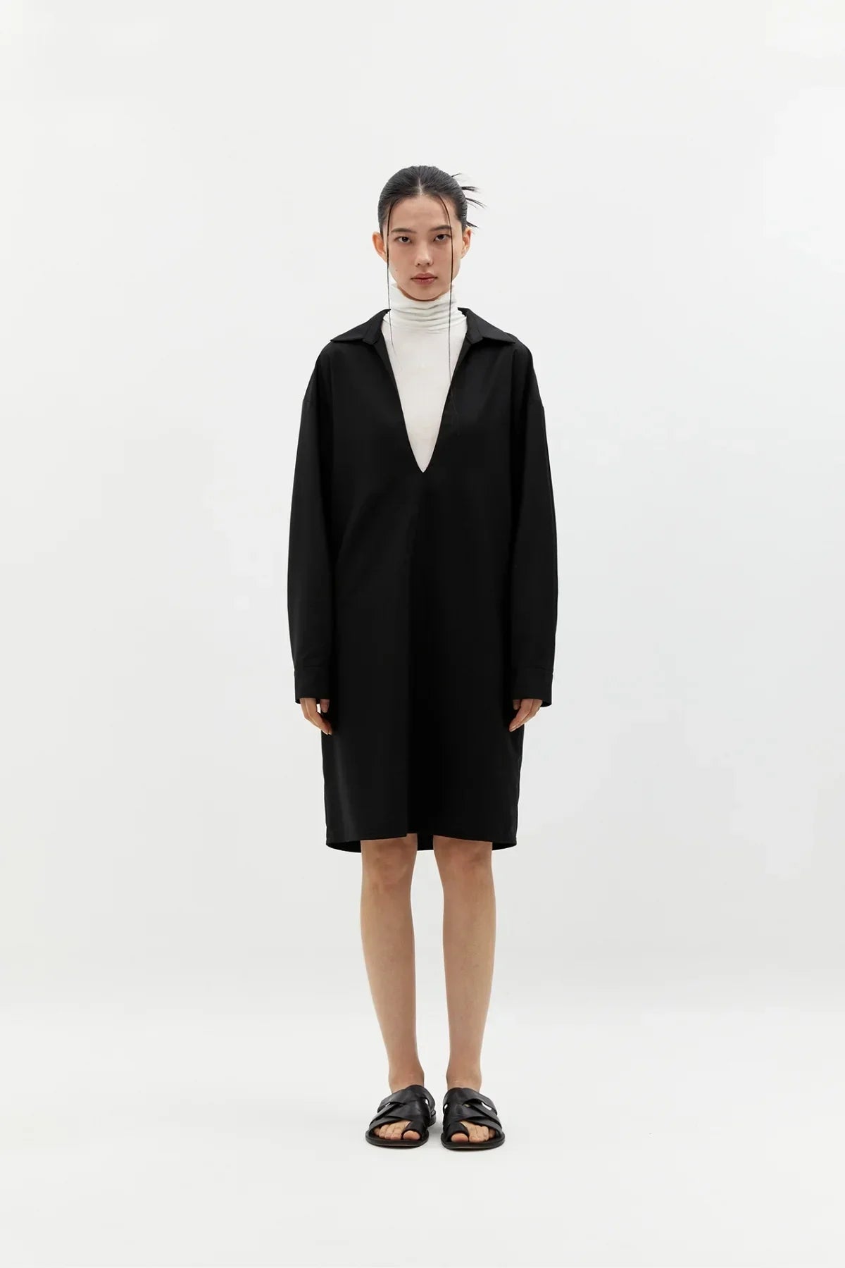 Model wearing black V-neck long dress over white turtleneck, paired with black sandals, white studio background