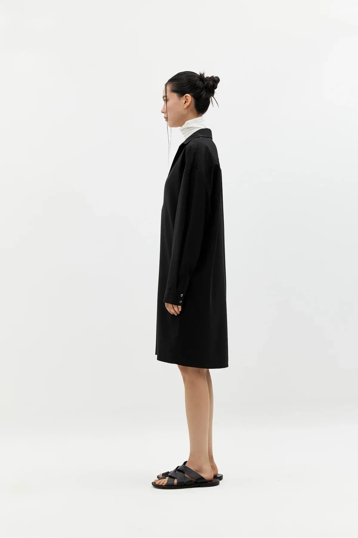 Side view of woman wearing black long-sleeve V-neck dress with white turtleneck and black sandals on white background