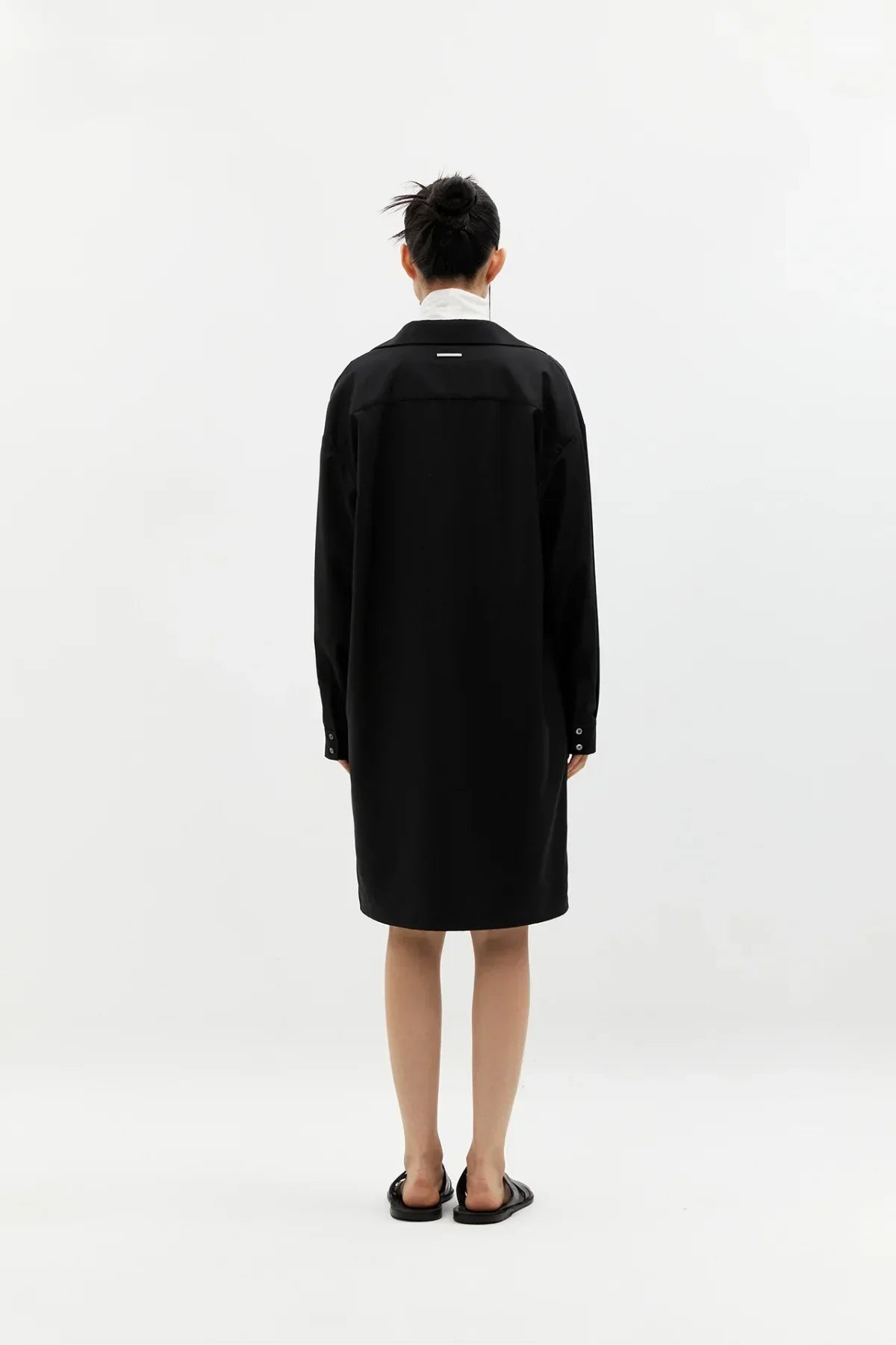Back view of a person wearing a black long sleeve knee-length coat with black sandals on white background