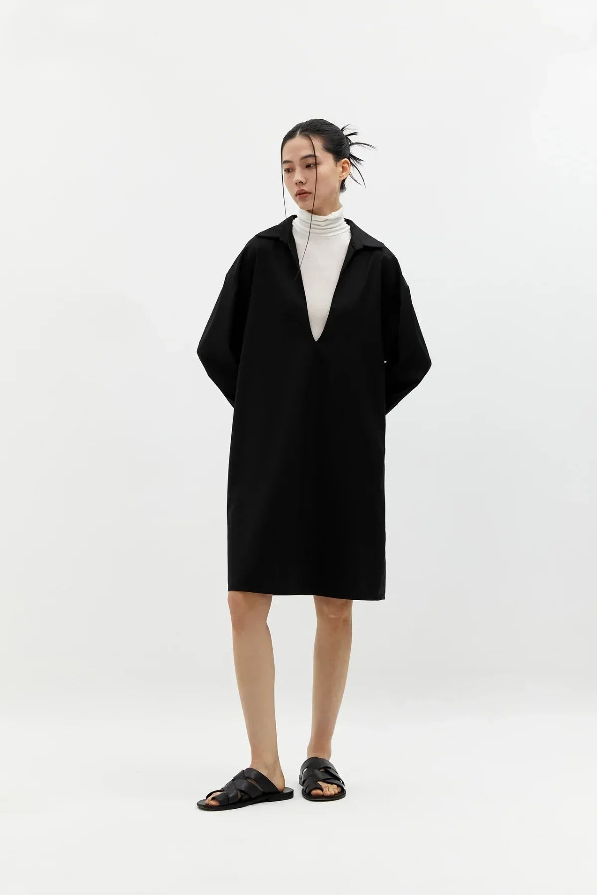 Woman wearing black V-neck long dress over white turtleneck with black sandals in studio