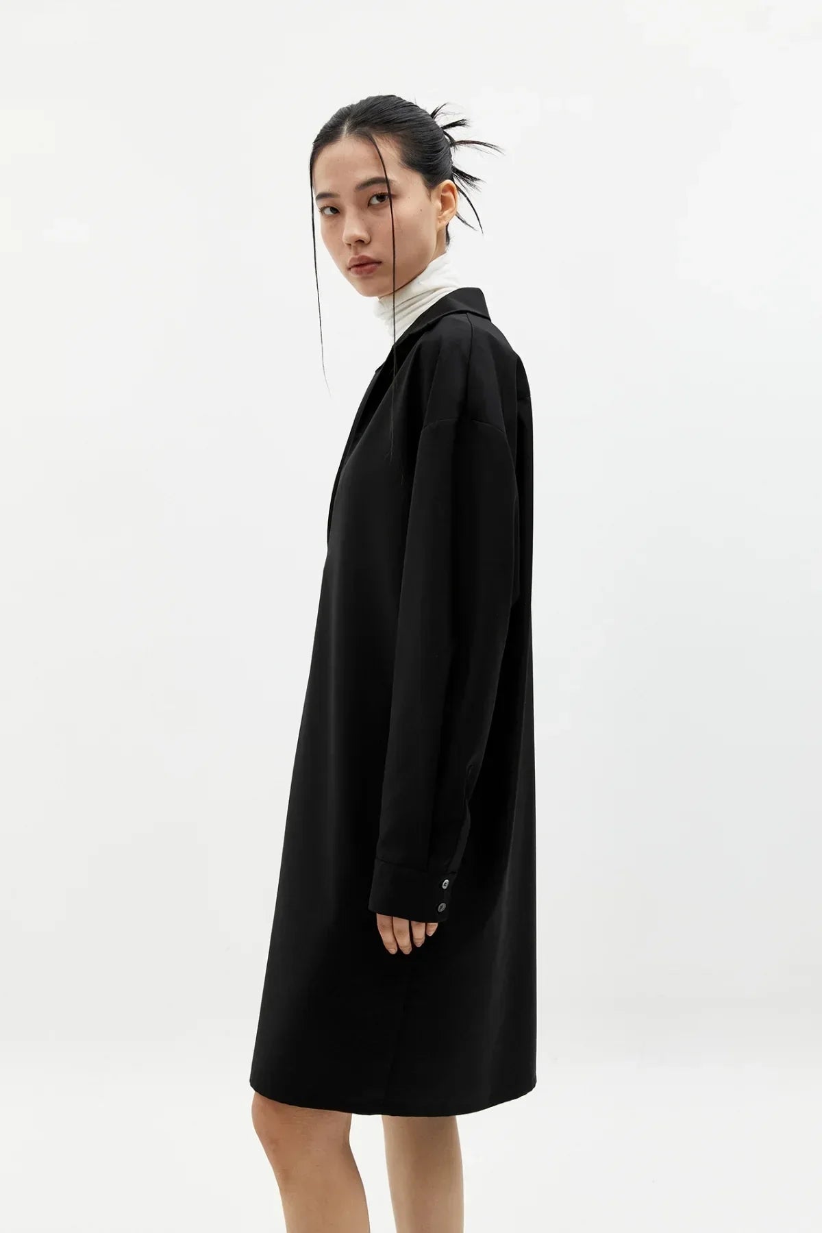 Model wearing black oversized long sleeve V-neck dress with white turtleneck, minimal studio background