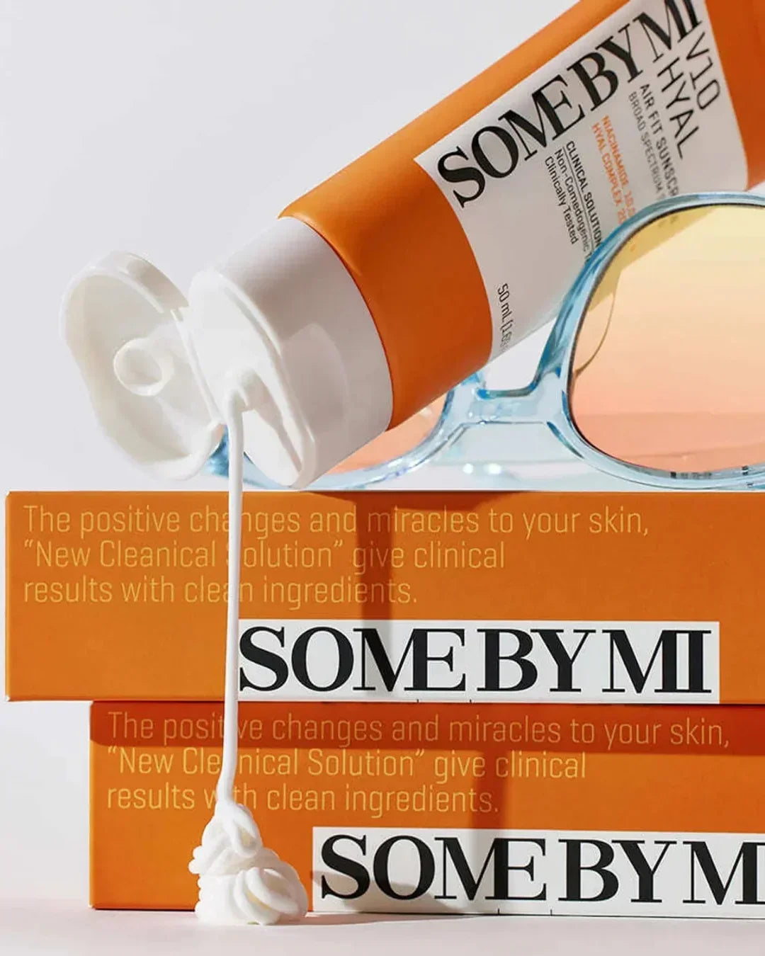 Some By Mi V10 Hyal Air Fit Sunscreen tube squeezing cream over orange product boxes with sunglasses