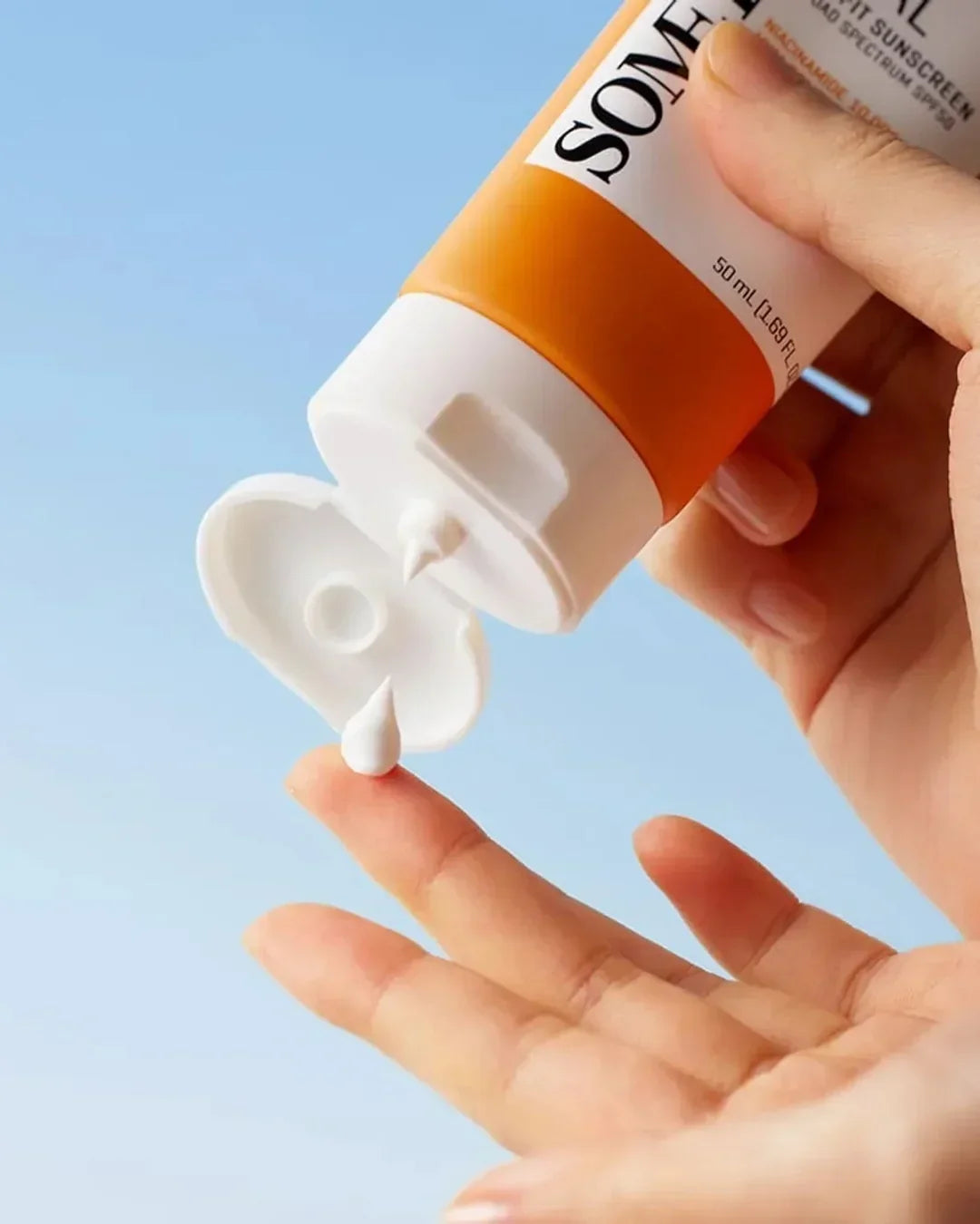 Hand squeezing white sunscreen cream from orange and white SOMEL tube against blue sky background