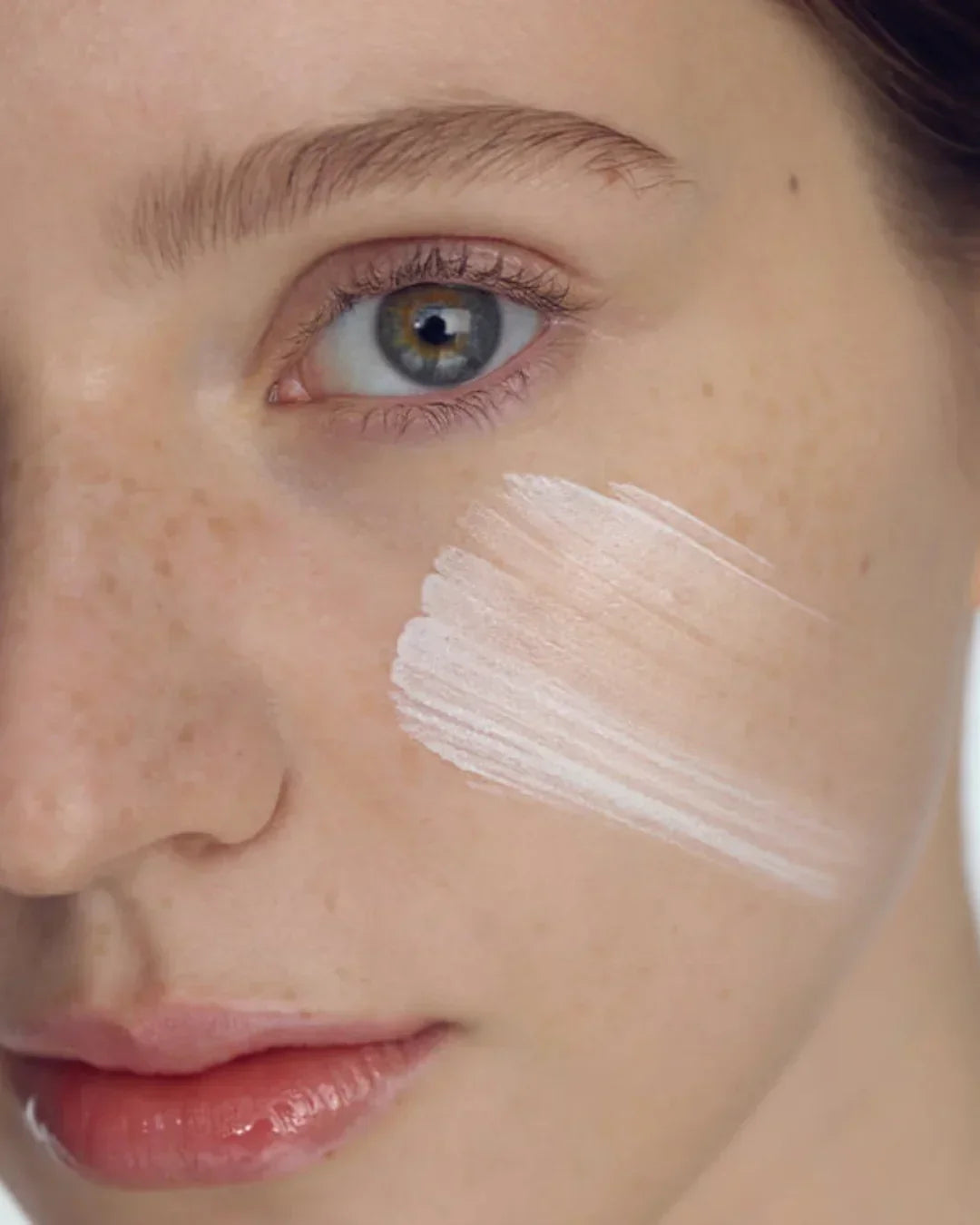 Close-up of a woman's face with fair skin and freckles, applying white sunscreen cream on her cheek