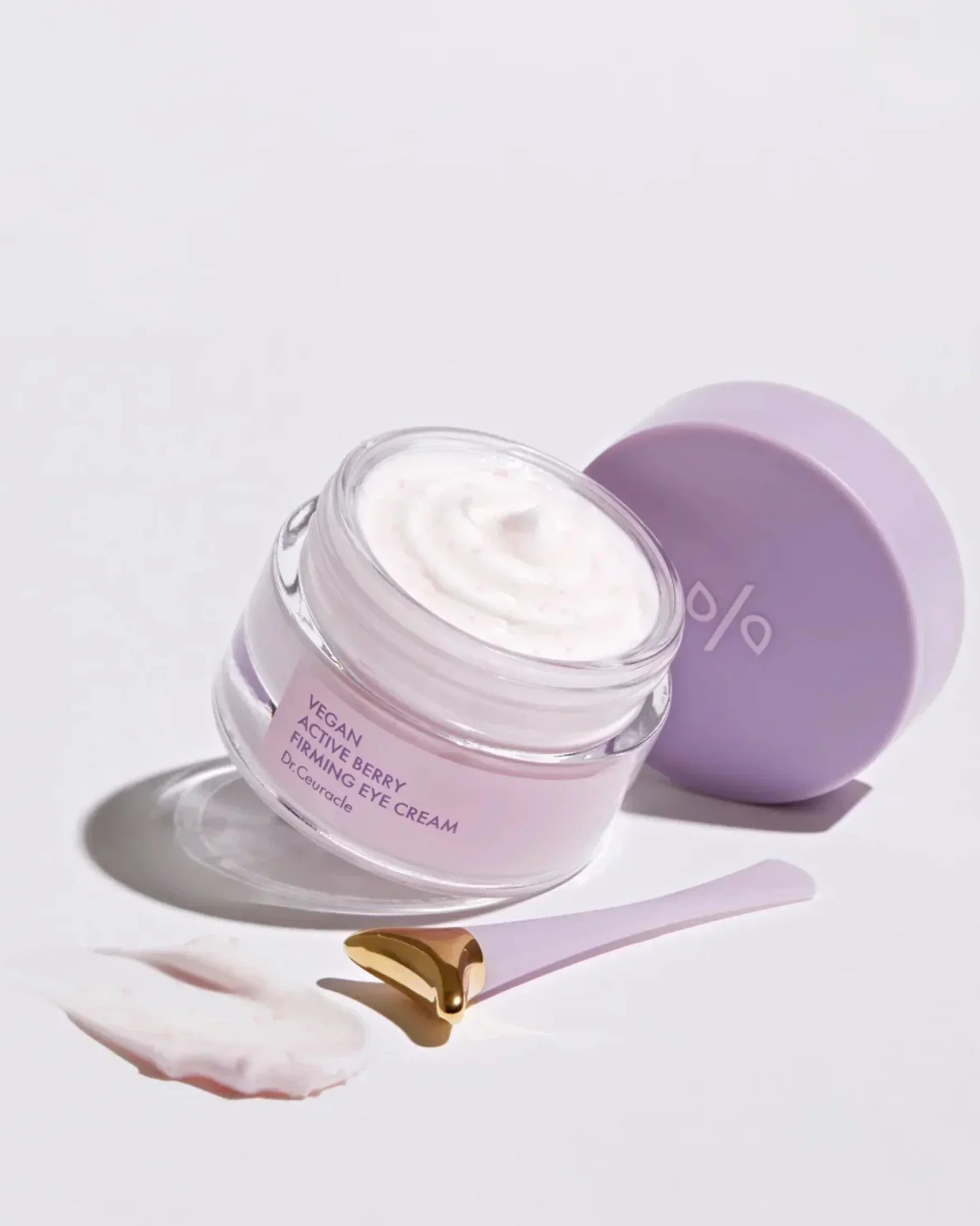 Open jar of vegan active berry firming eye cream with lavender lid and gold-tipped applicator on white background