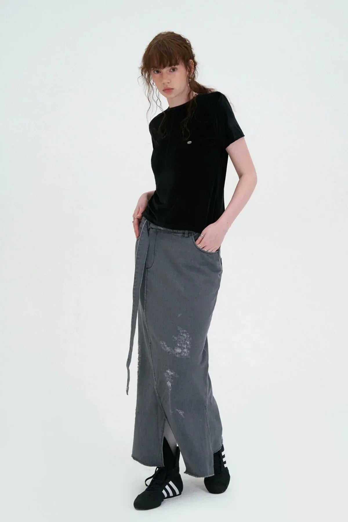Young woman wearing black velvet T-shirt and distressed grey long skirt with black sneakers on white background