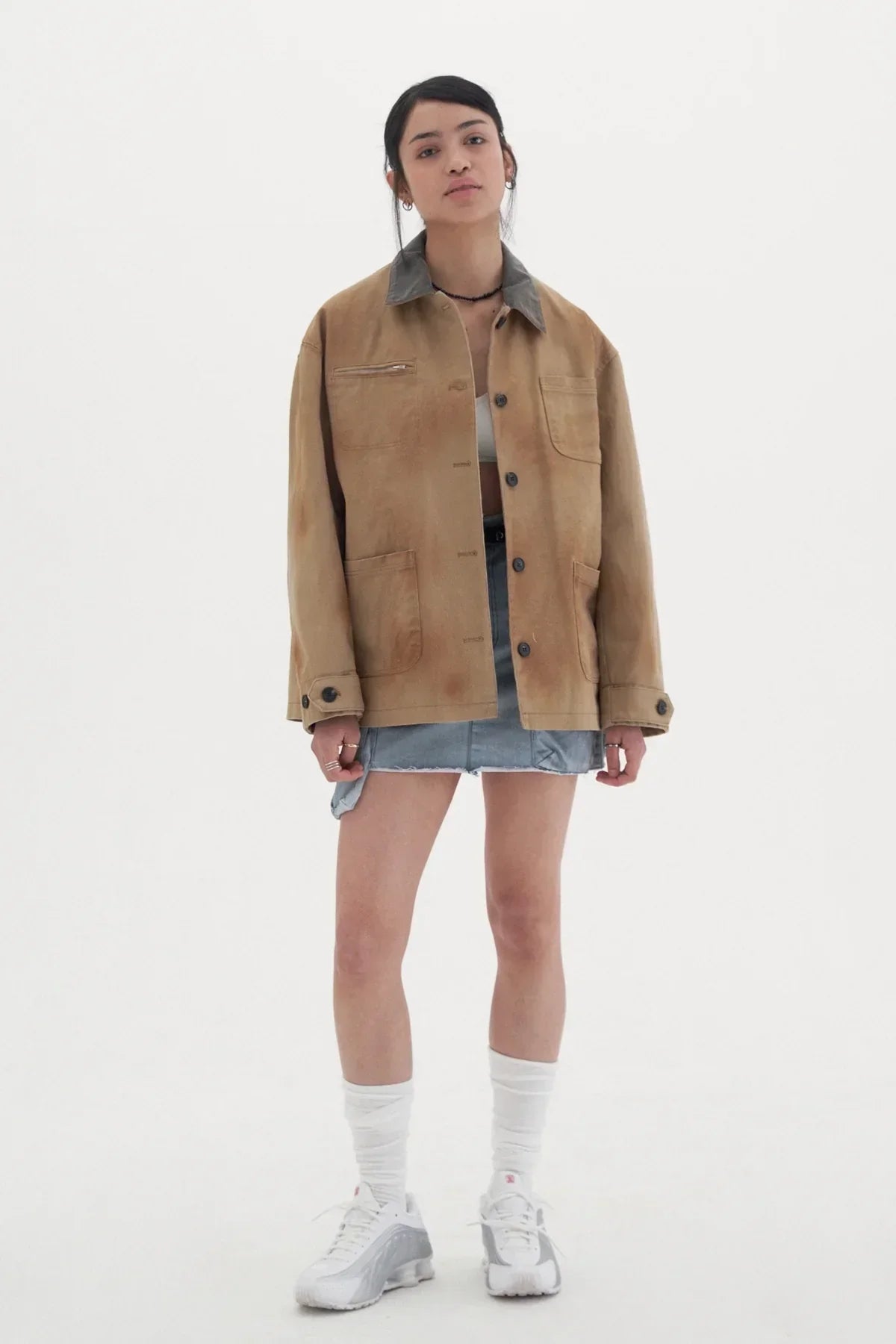 Young woman wearing oversized beige vintage work jacket with dark collar, denim skirt, white socks, and sneakers