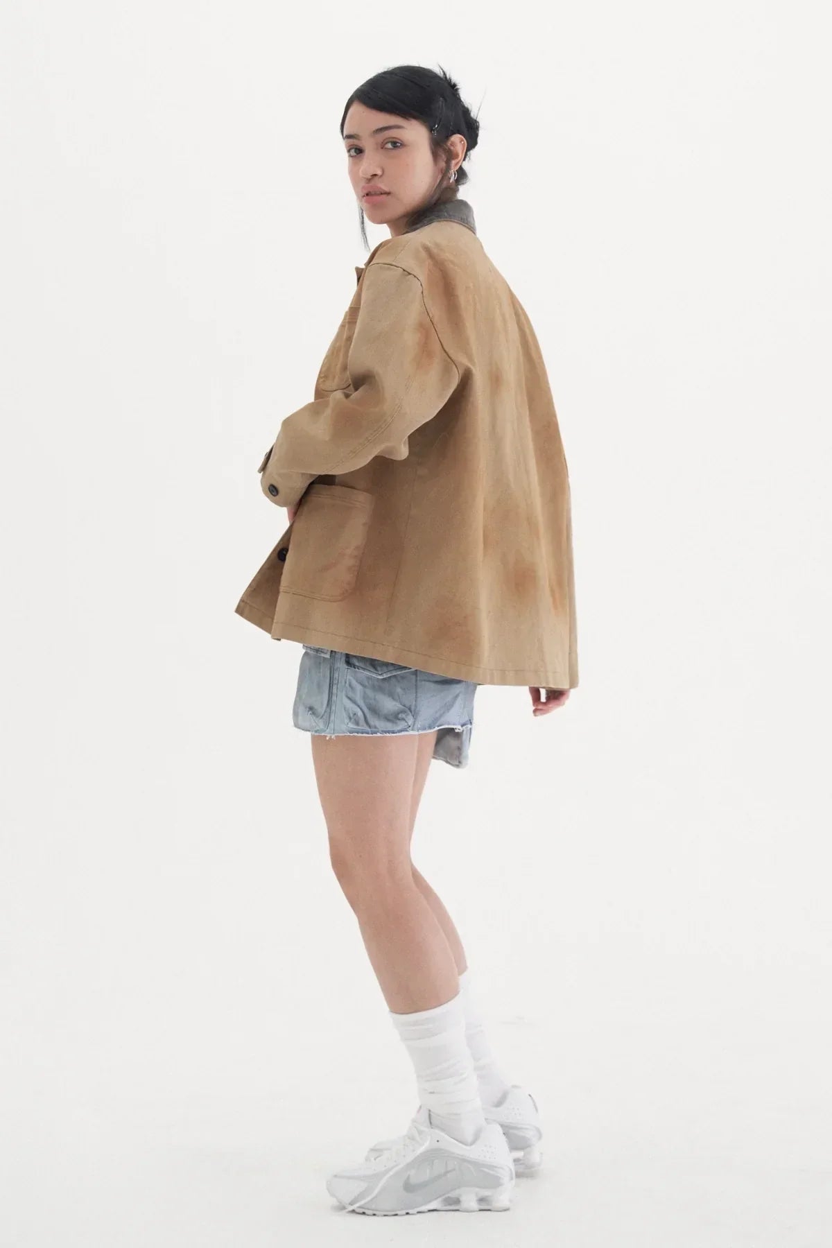 Young woman wearing a vintage beige work jacket with denim shorts and white sneakers on white background