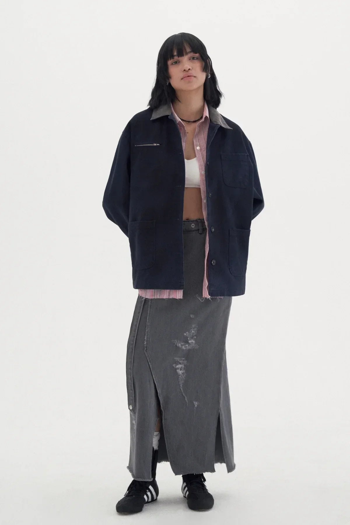 Young woman wearing a navy vintage work jacket over a pink shirt and white crop top, paired with a distressed grey maxi skirt and black sneakers