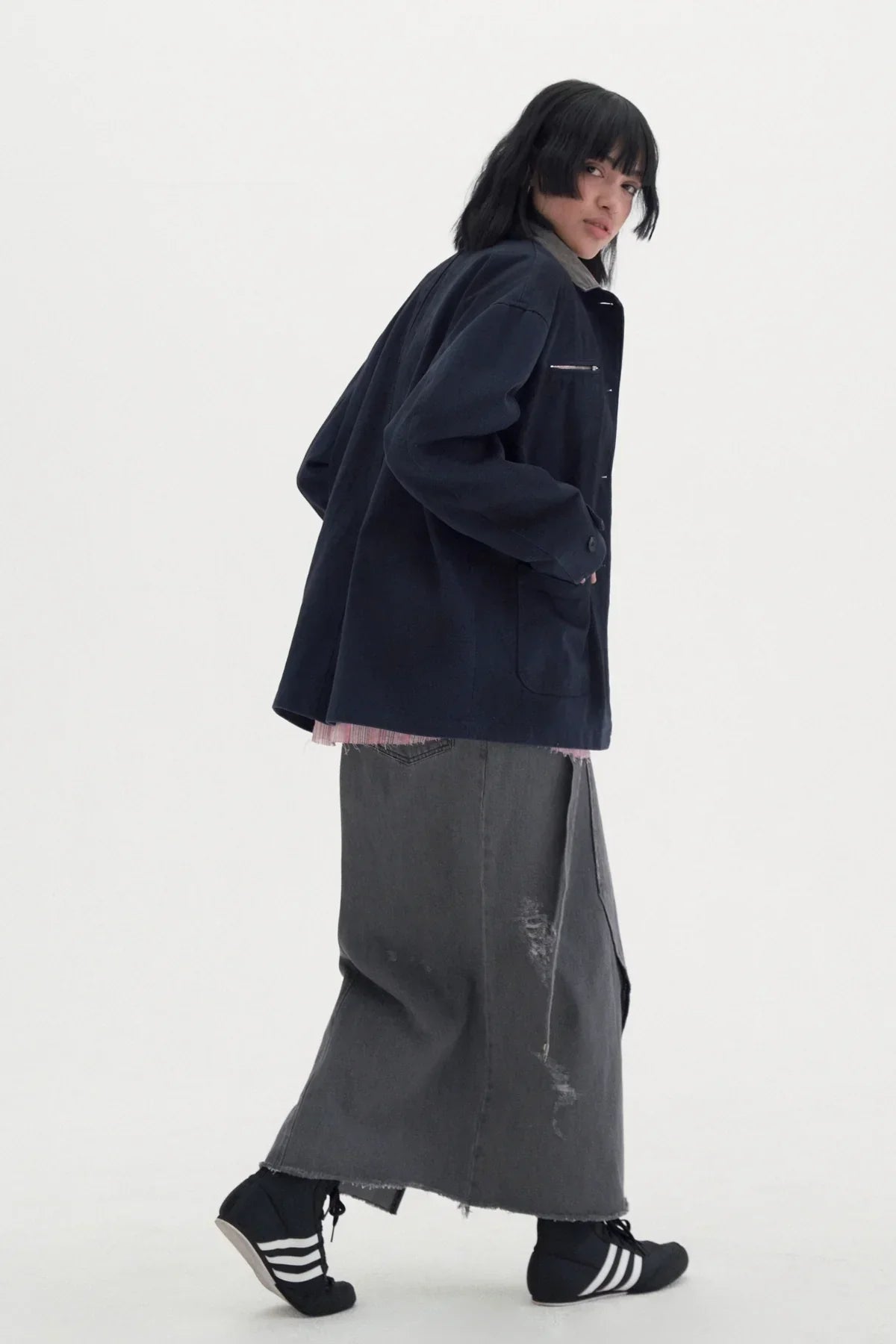 Model wearing navy vintage work jacket over pink shirt with gray distressed maxi skirt and black Adidas sneakers