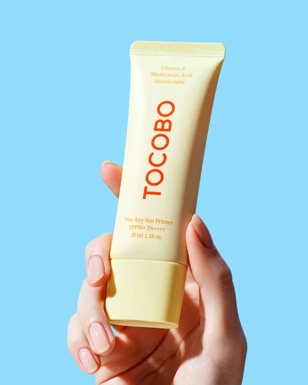 Hand holding a yellow tube of TOCOBO Vita Airy Sun Primer SPF50+ PA++++ sunscreen against a blue background