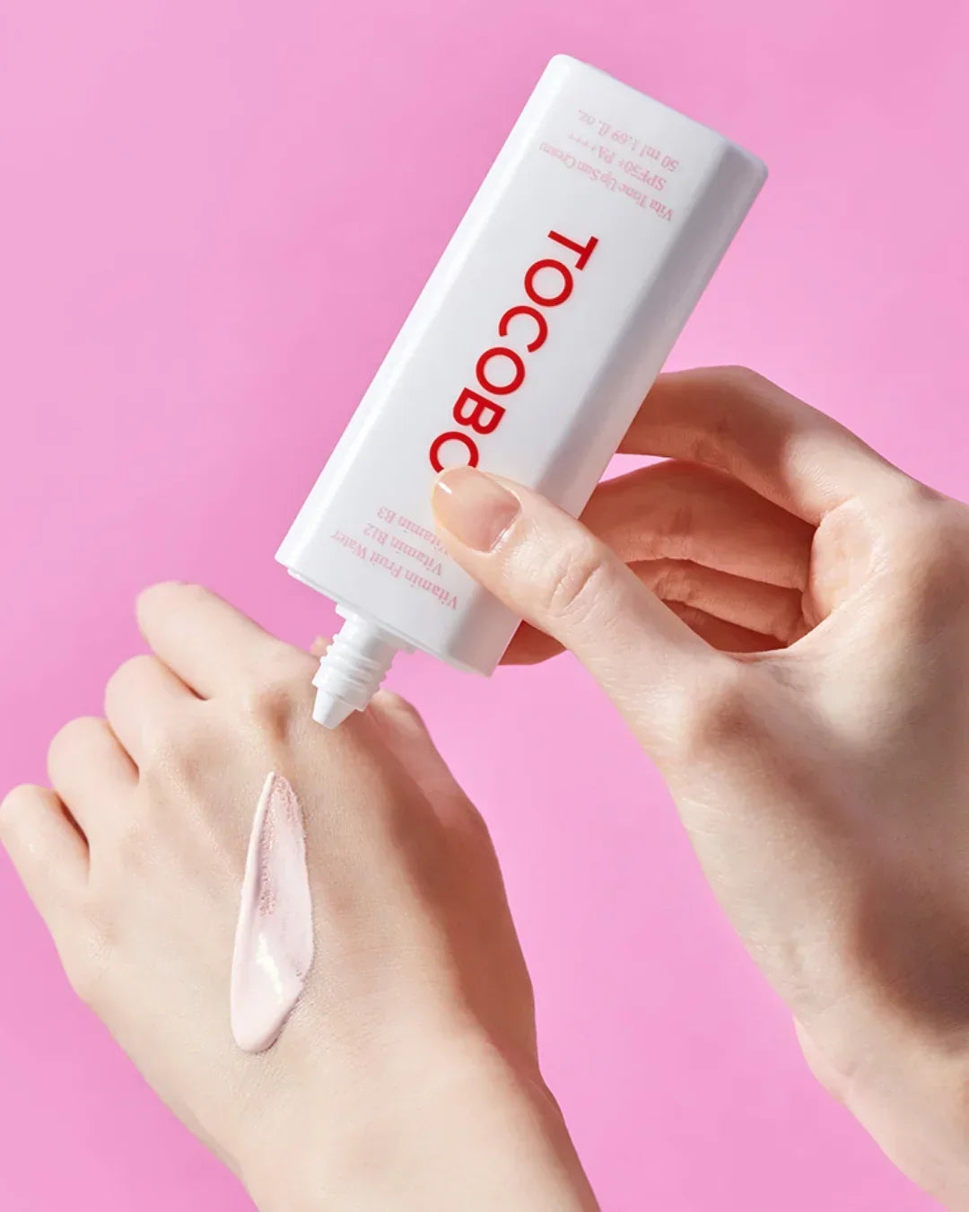 Person applying light pink TOCOBOO Vita Tone Up Sun Cream SPF50 PA++ on hand against pink background