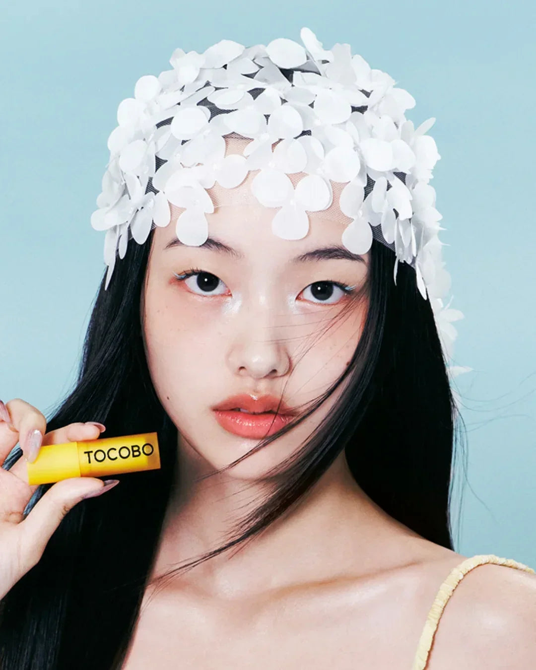 Young woman wearing white floral swim cap, holding yellow TOCOBO lip balm against light blue background