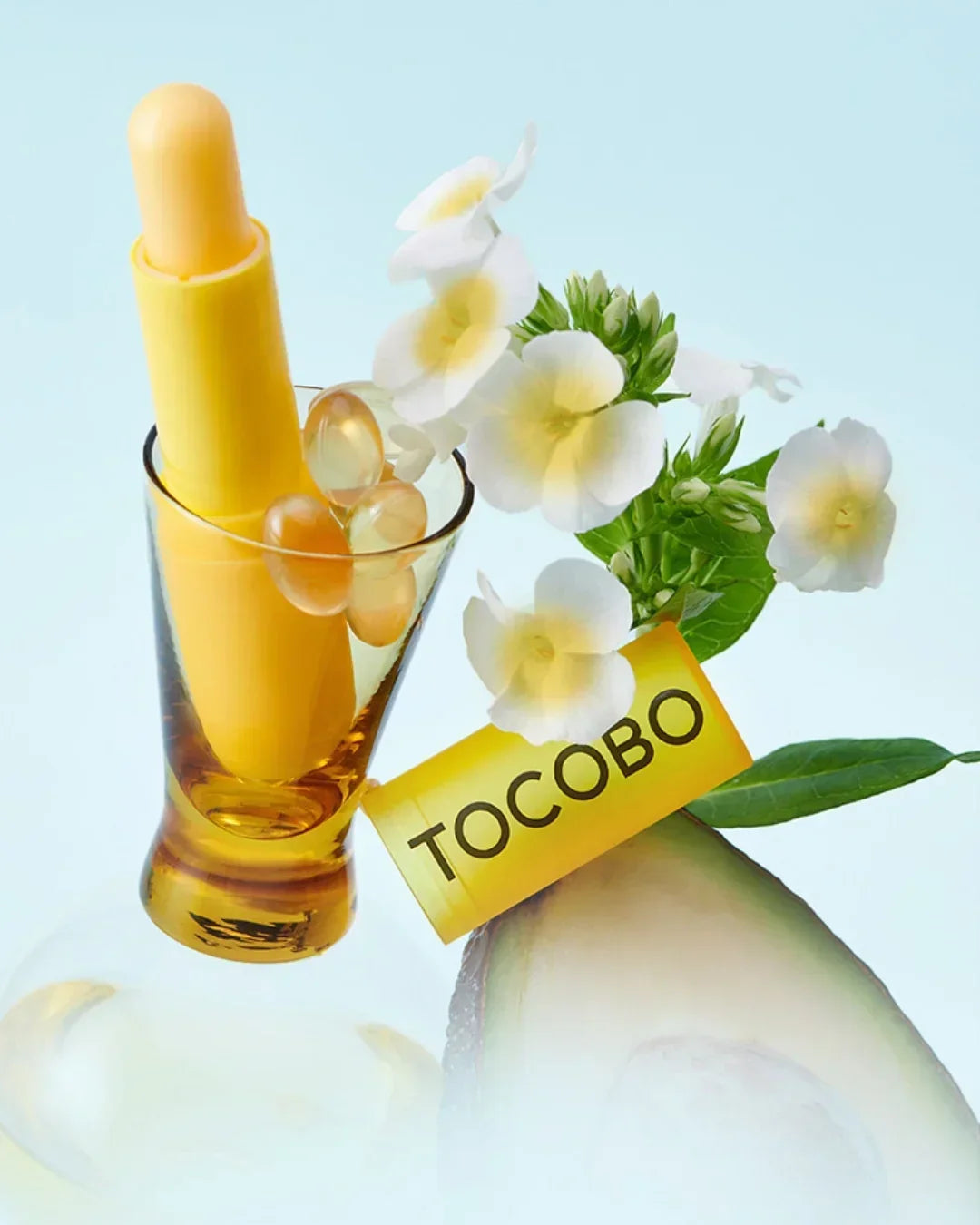 Yellow TOCOBO vitamin nourishing lip balm with capsules, white flowers, and avocado slice on light blue background