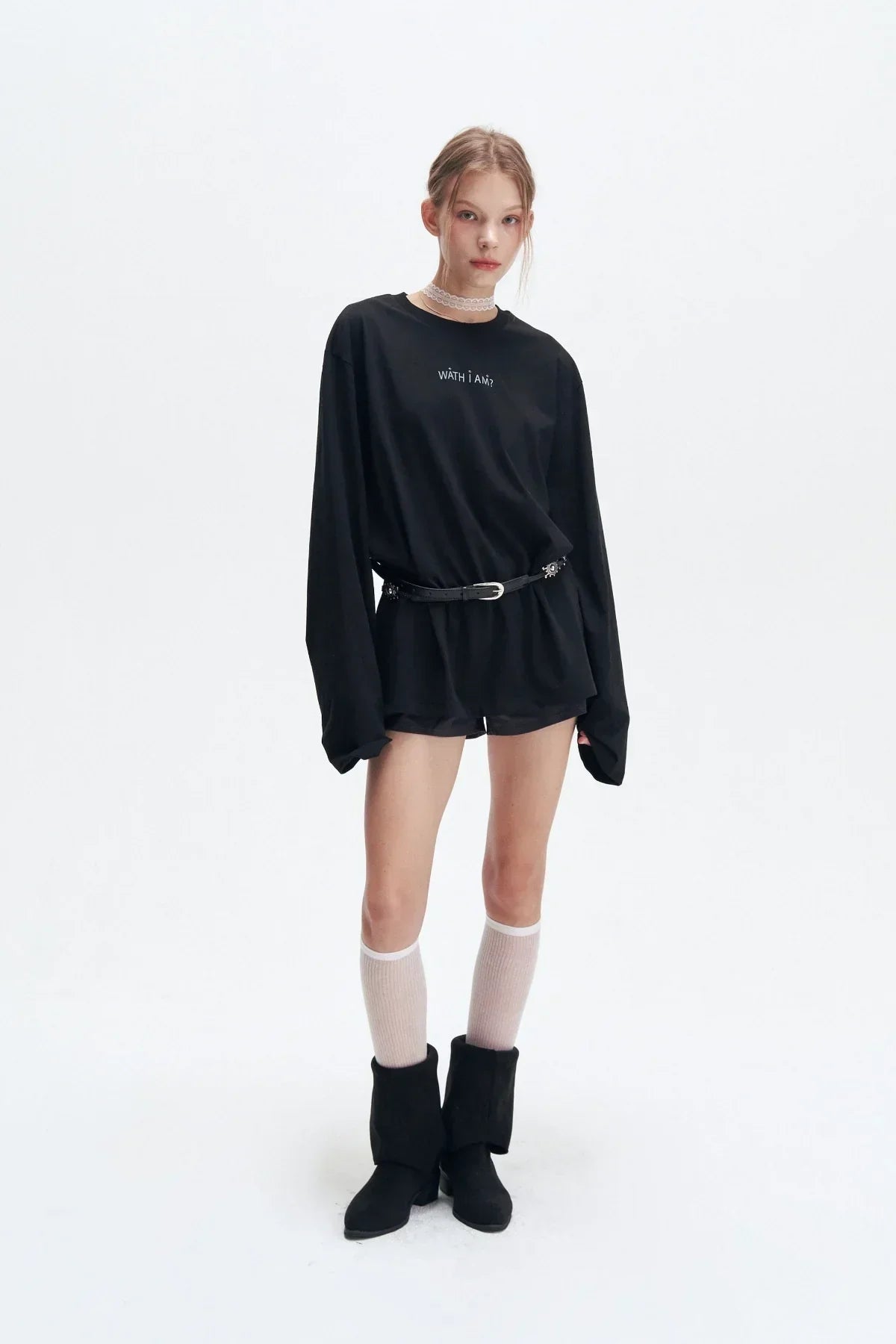 Young woman in black oversized long sleeve top with text, black belt, shorts, white knee socks, and black boots