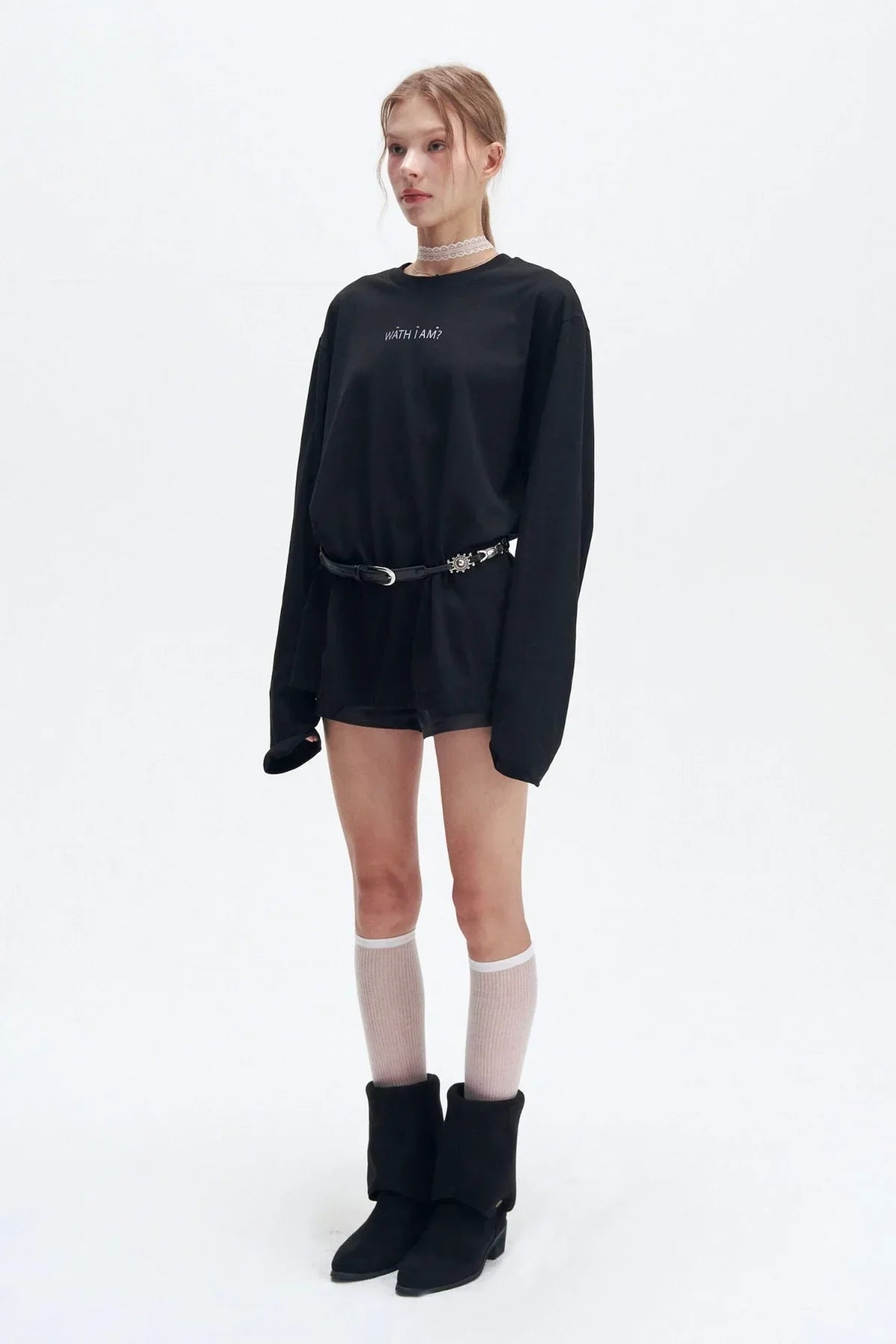 Young woman wearing oversized black long-sleeve shirt with text, black belt, knee-high white socks, and black boots on white background