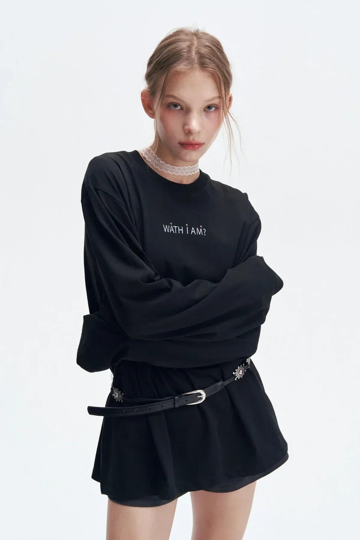 Young woman wearing black long sleeve shirt with 'WATH I AM?' text, black skirt, belt, and white lace choker