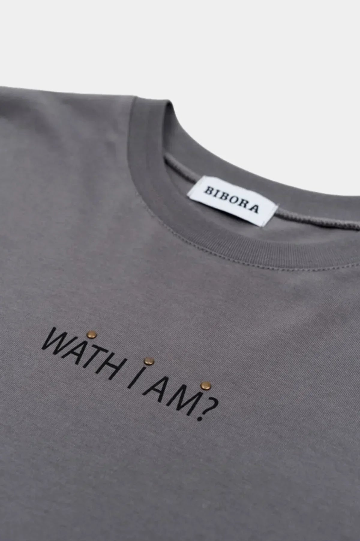 Close-up of grey long sleeve shirt with 'WATH I AM?' text and round metallic embellishments near neckline