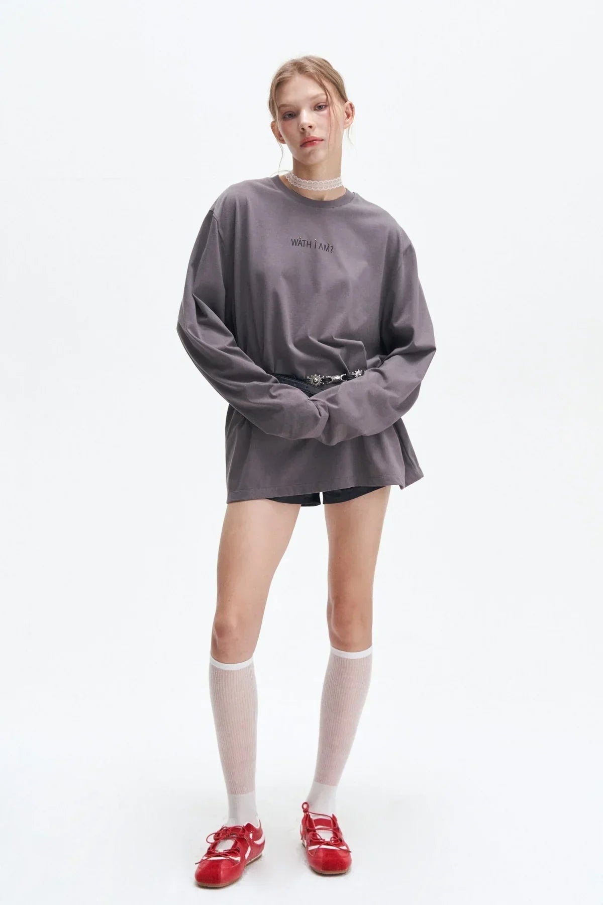 Young woman wearing oversized gray long-sleeve shirt with text, black shorts, white knee-high socks, and red shoes