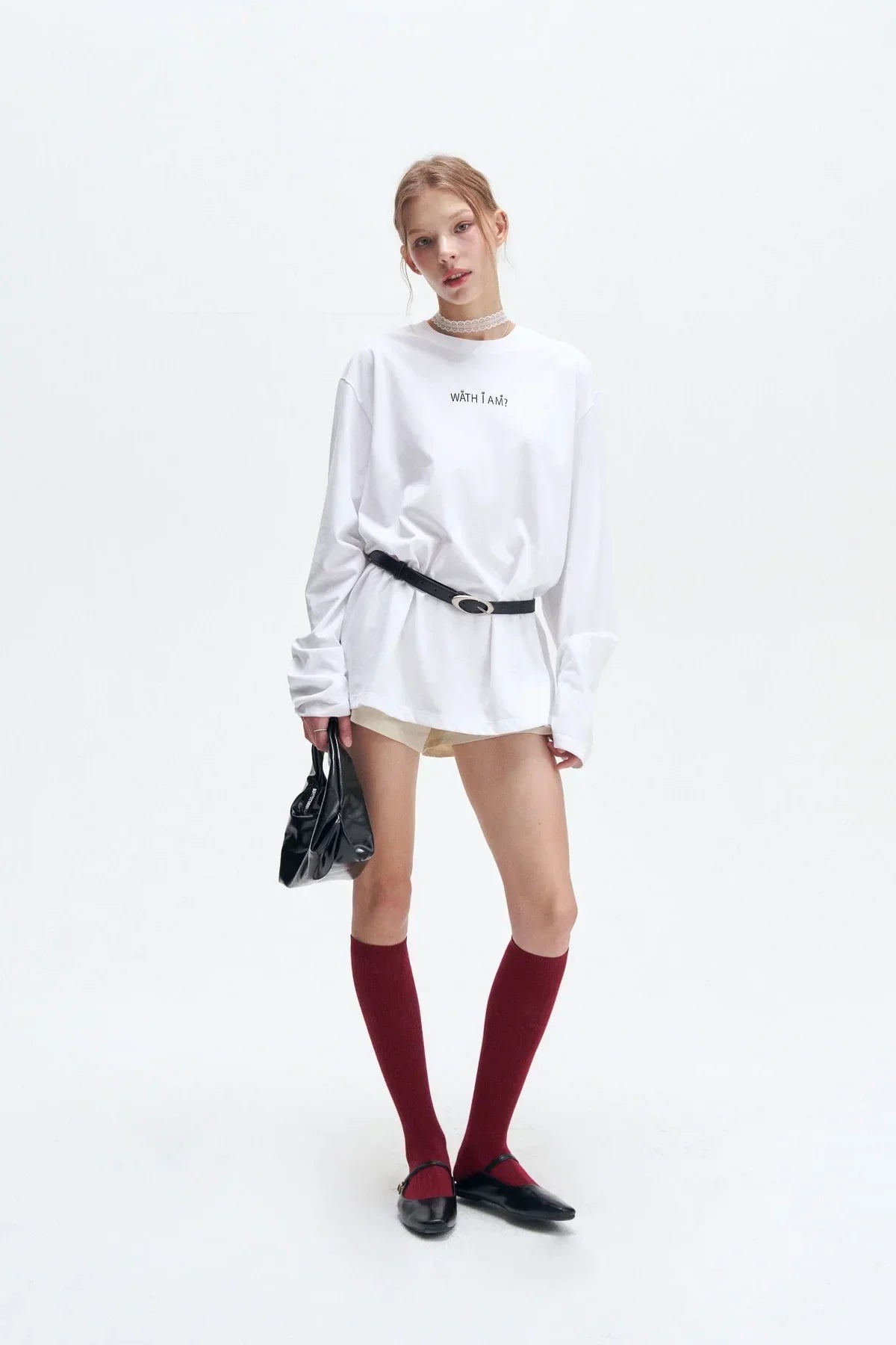 Model wearing white long-sleeve oversized shirt with text, black belt, burgundy knee-high socks, black flats, and holding black handbag