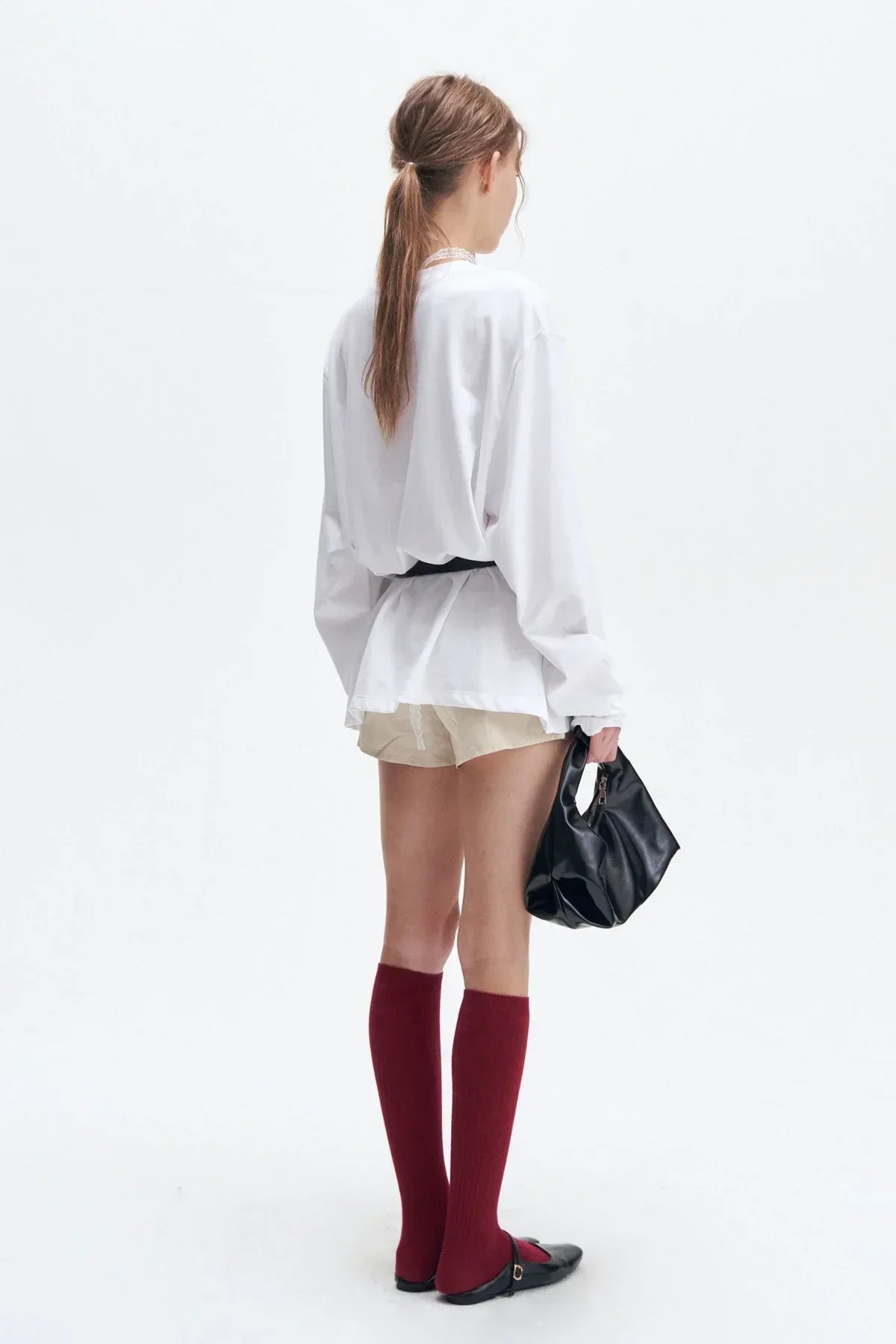 Woman wearing white long sleeve shirt with black belt, beige shorts, red knee-high socks, black flats, holding black handbag