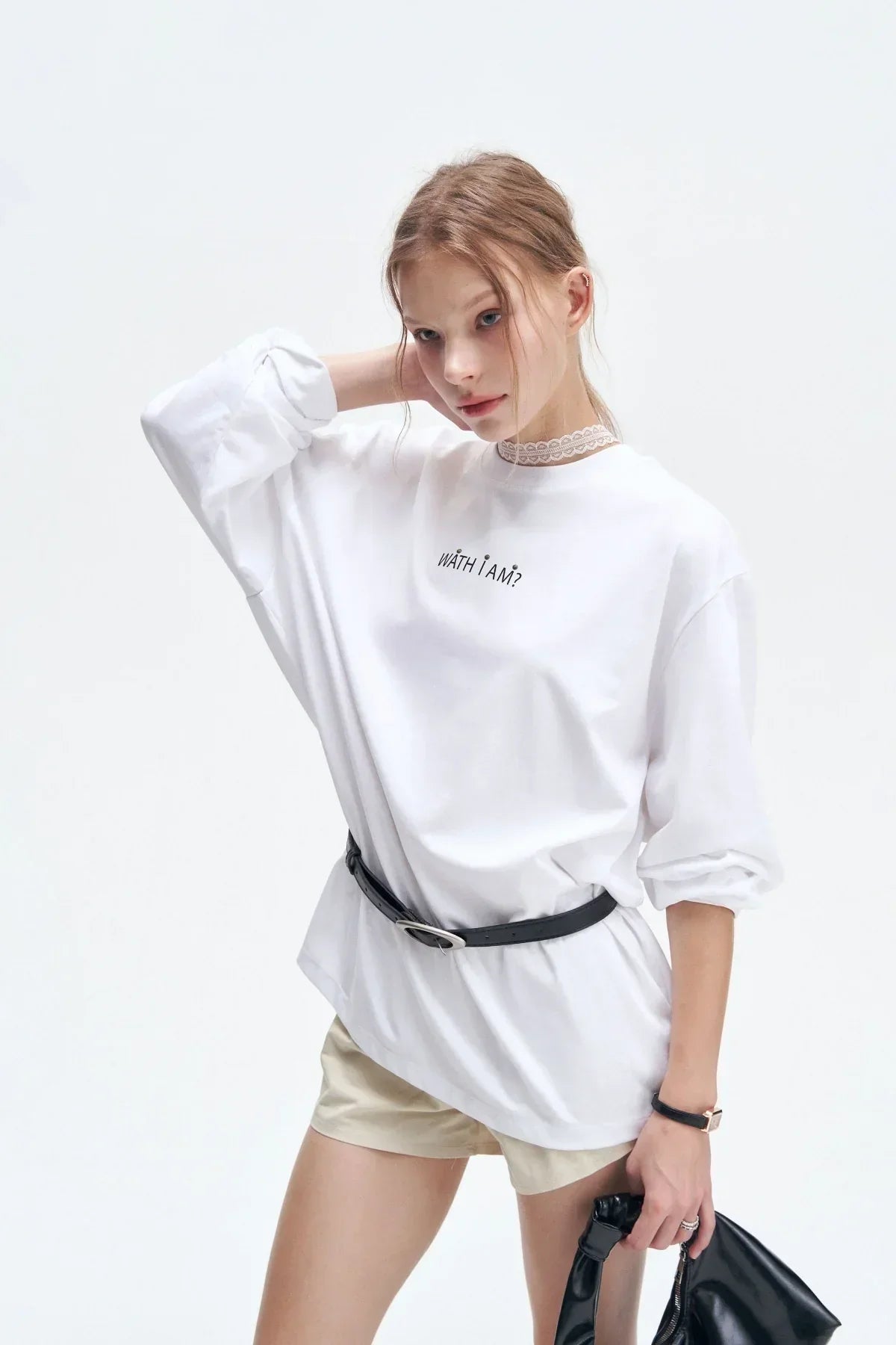 Young woman in white long-sleeve shirt with text, beige shorts, black belt, and holding black handbag