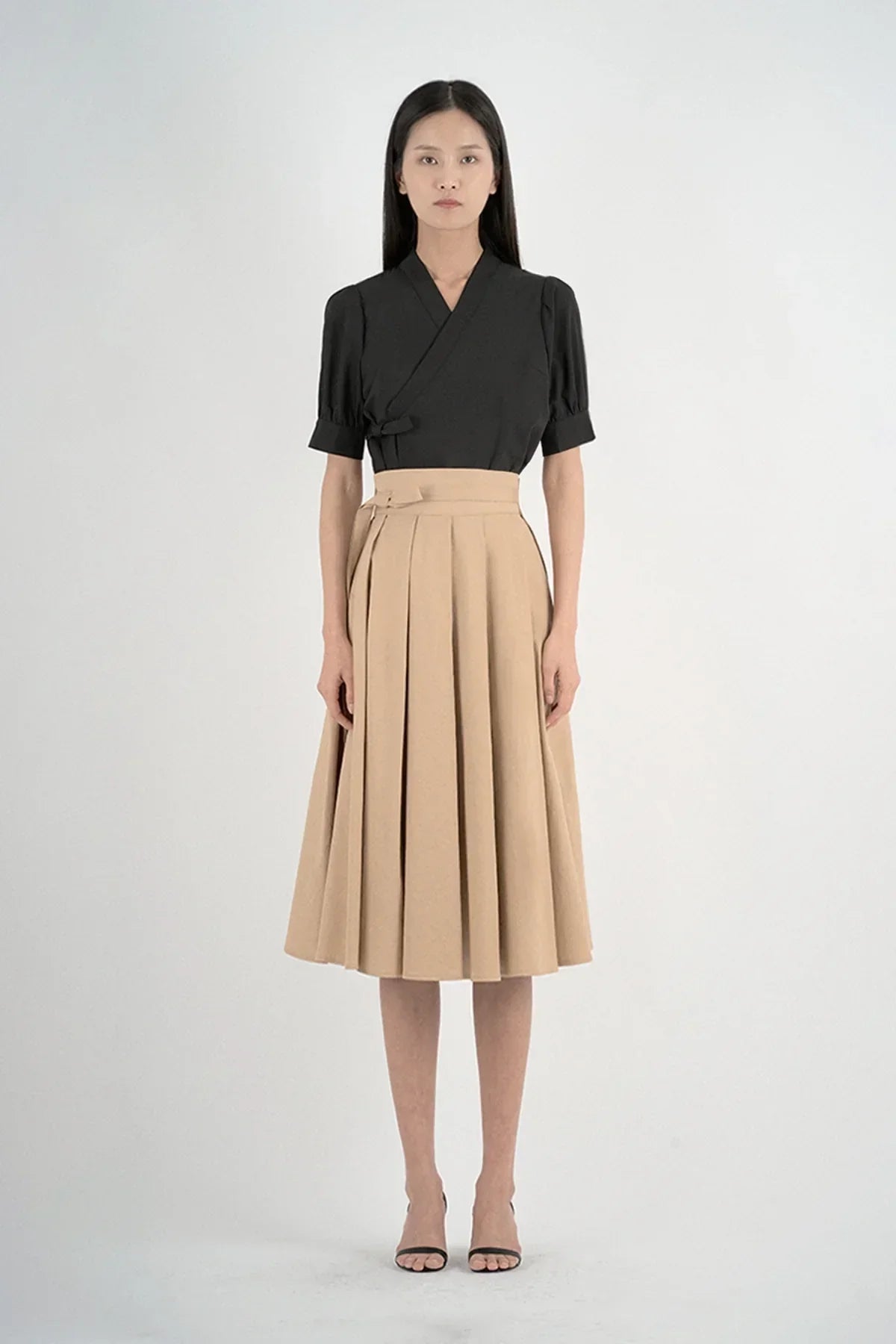 Woman wearing a black short-sleeve top and a beige high-waisted pleated midi skirt with black heels
