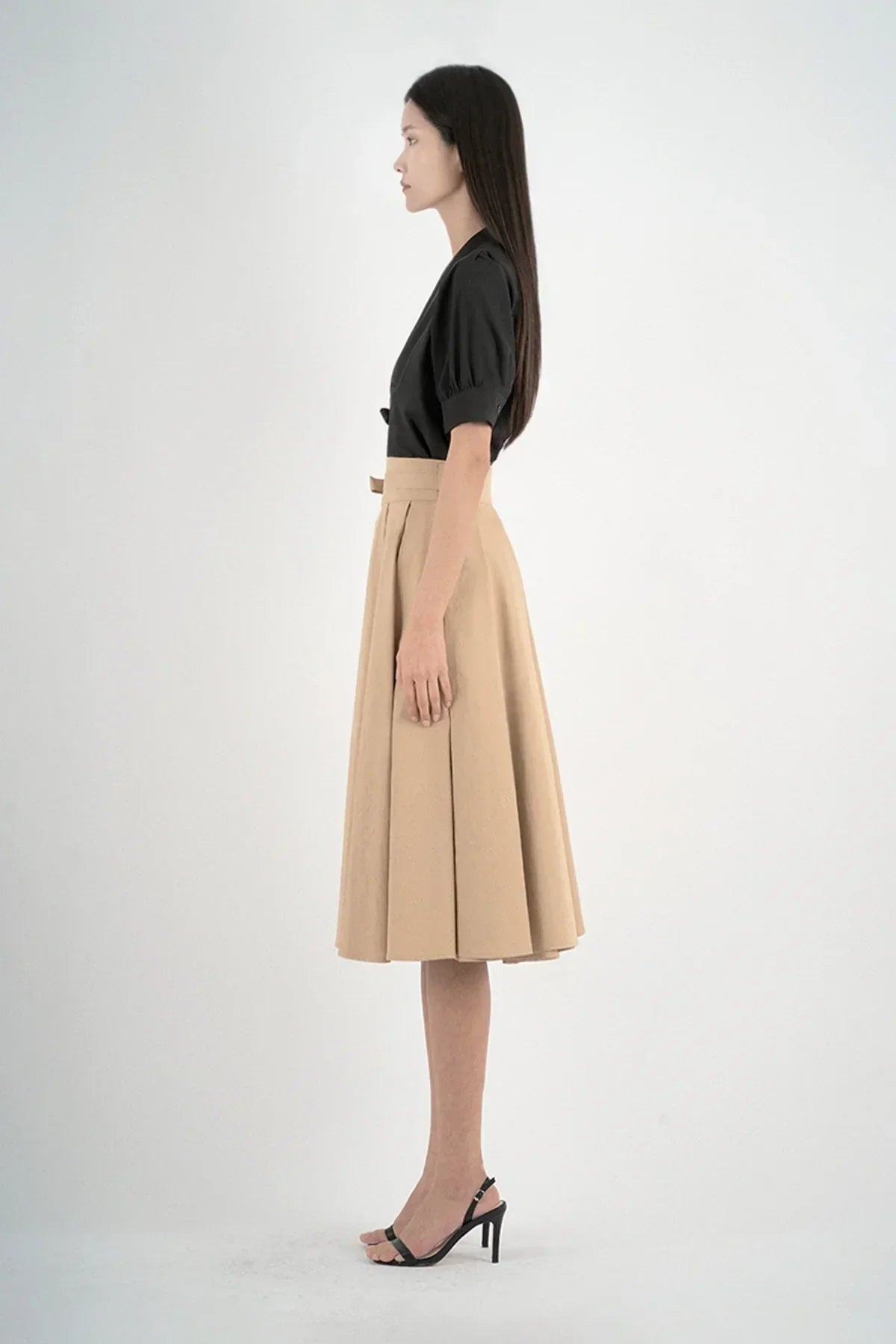 Side view of woman in black blouse and beige midi skirt with pleats and black high heel sandals