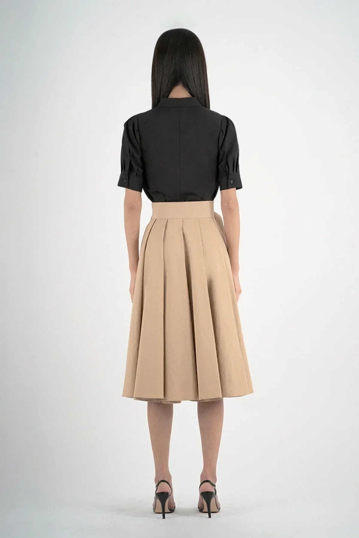 Back view of woman wearing black shirt and beige pleated midi skirt with black high heels