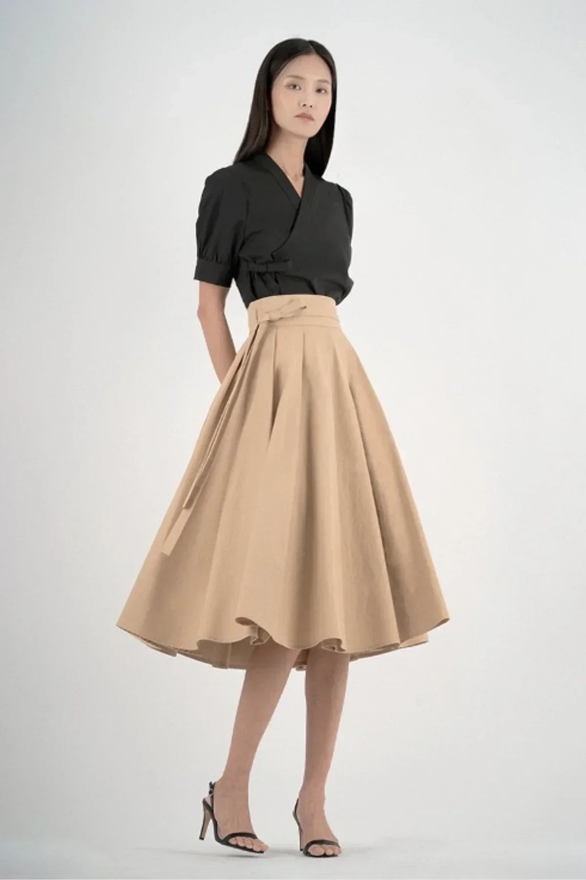 Woman wearing black wrap top and beige high-waisted A-line midi skirt with bow detail in studio