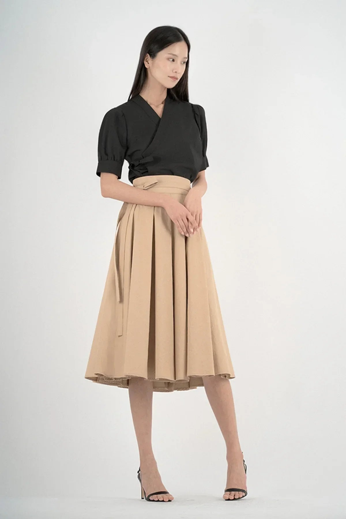 Woman wearing black puff sleeve top and beige pleated midi skirt with black high heels in studio