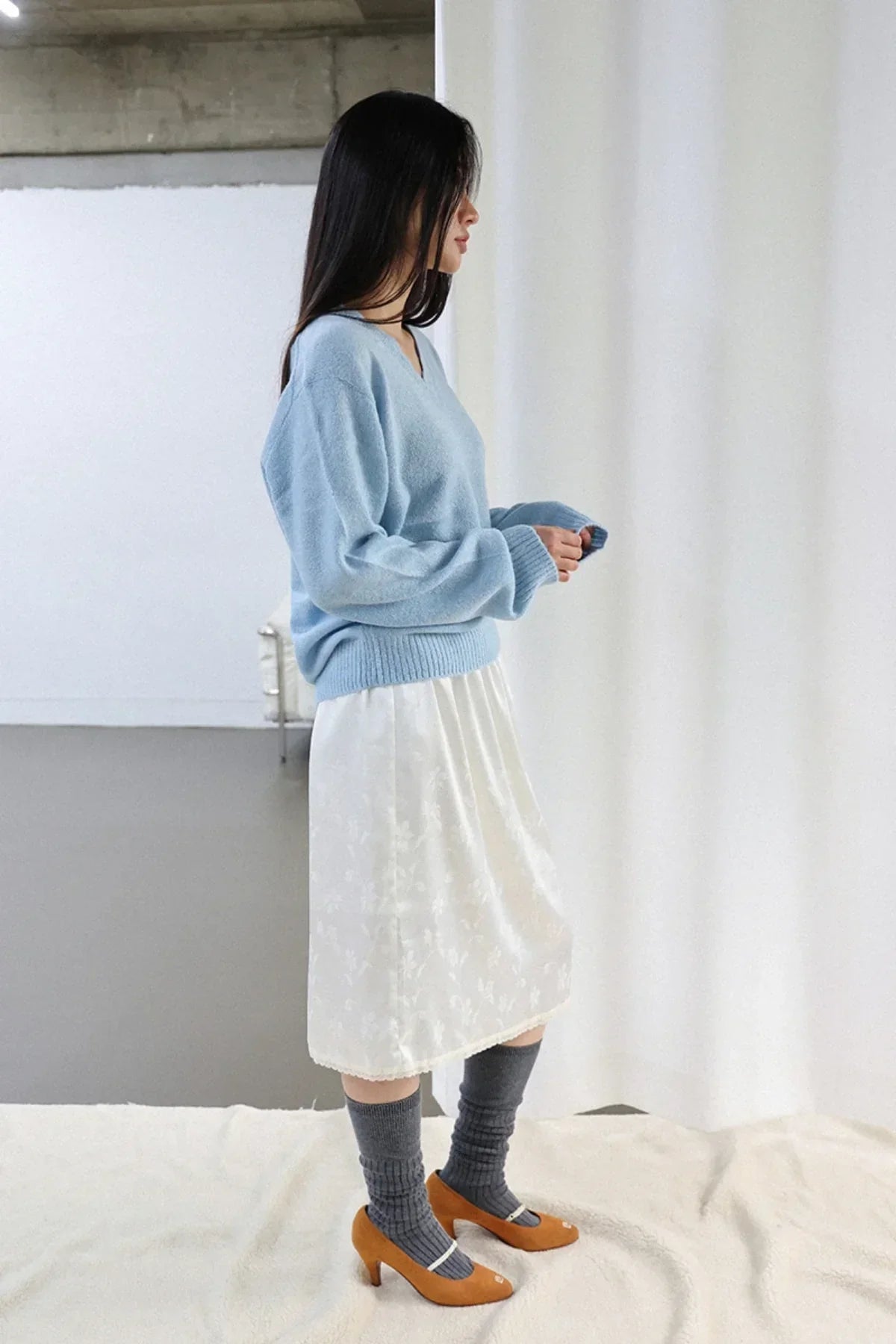 Woman wearing light blue V-neck alpaca knit sweater with white floral midi skirt, gray socks, and brown heels indoors