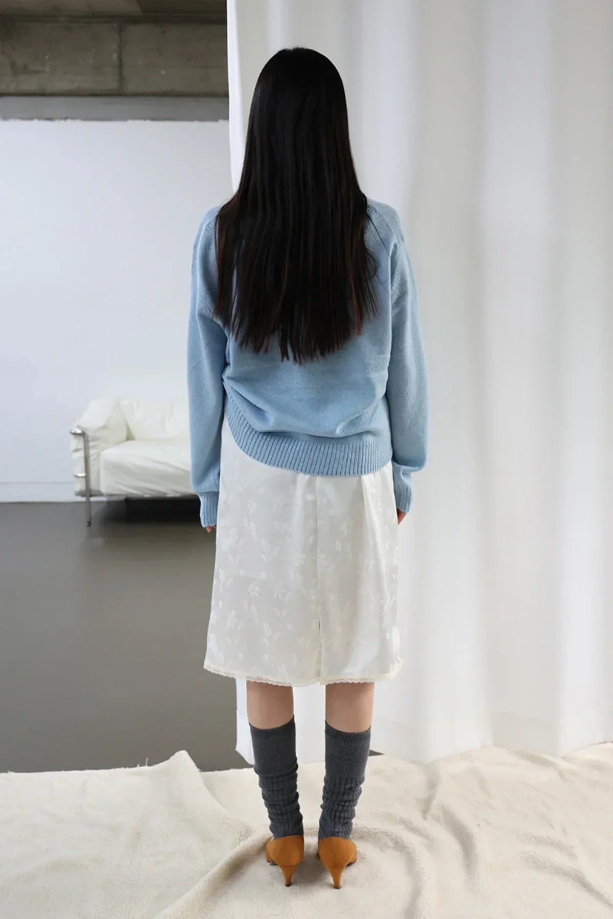 Woman with long black hair wearing a light blue sweater, white floral skirt, dark socks, and brown heels, standing indoors