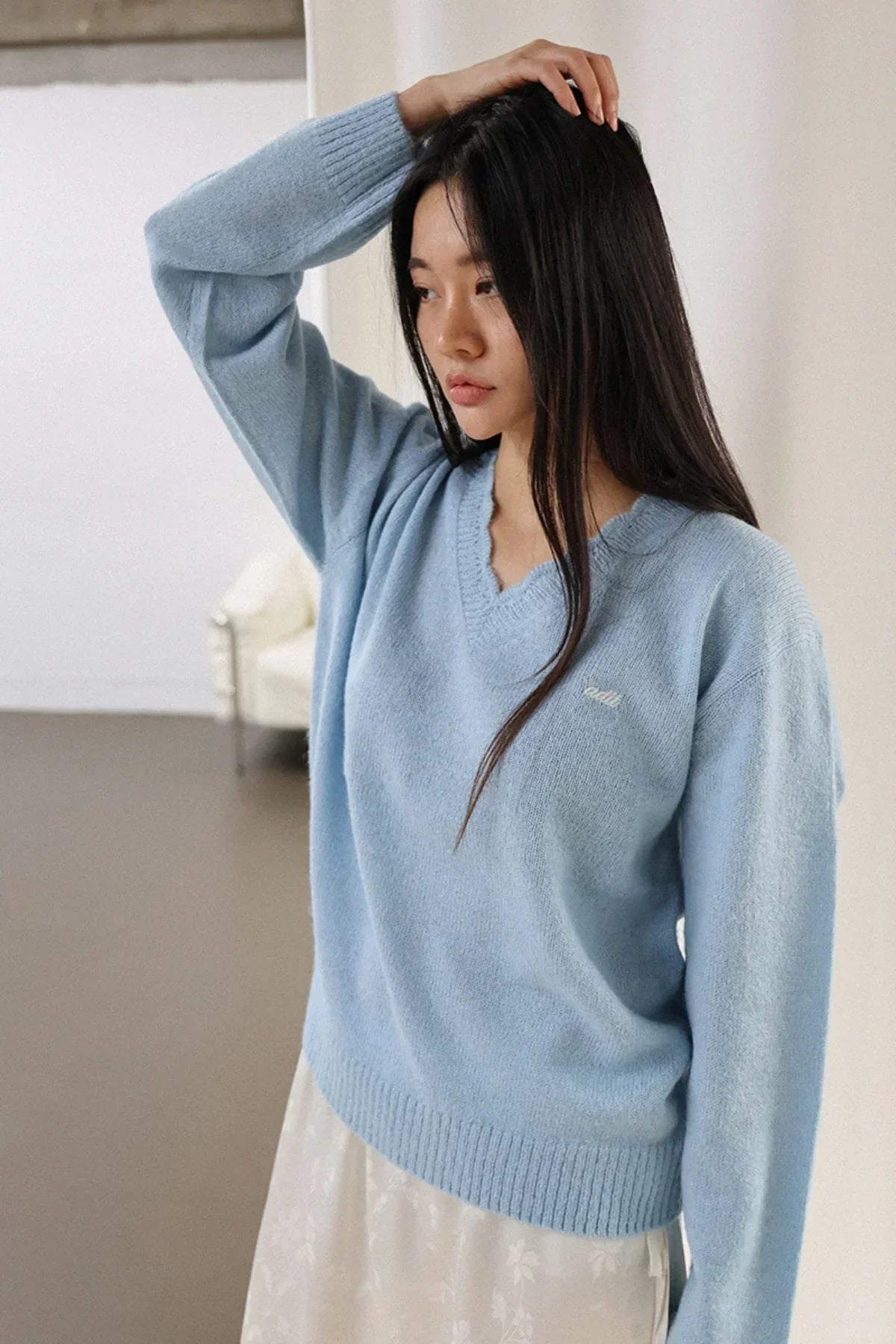 Woman wearing light blue alpaca knit V-neck sweater with white floral skirt in minimalist room