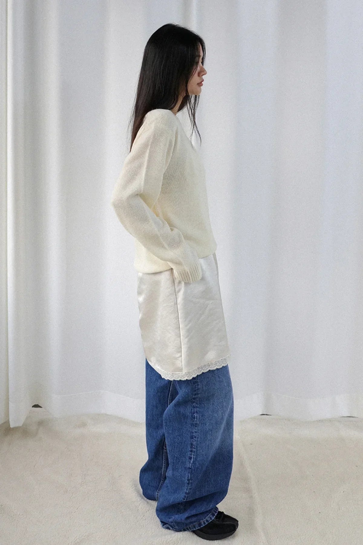 Side view of woman wearing ivory alpaca knit v-neck sweater, ivory satin slip skirt, loose blue jeans, black shoes