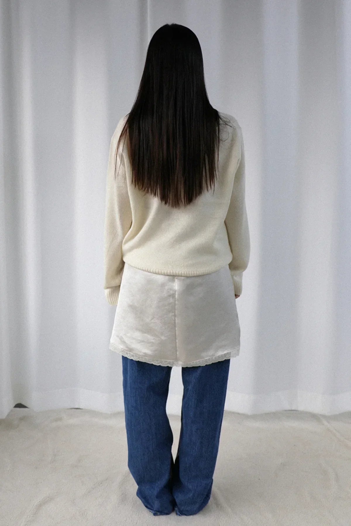 Woman with long dark hair wearing ivory alpaca knit sweater, cream skirt, and blue jeans standing indoors