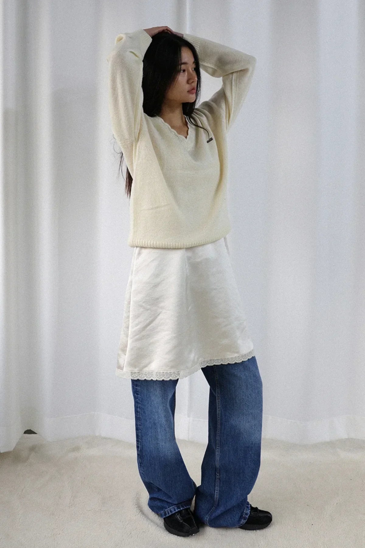 Woman wearing ivory alpaca knit V-neck sweater over white dress and blue jeans in minimalist setting