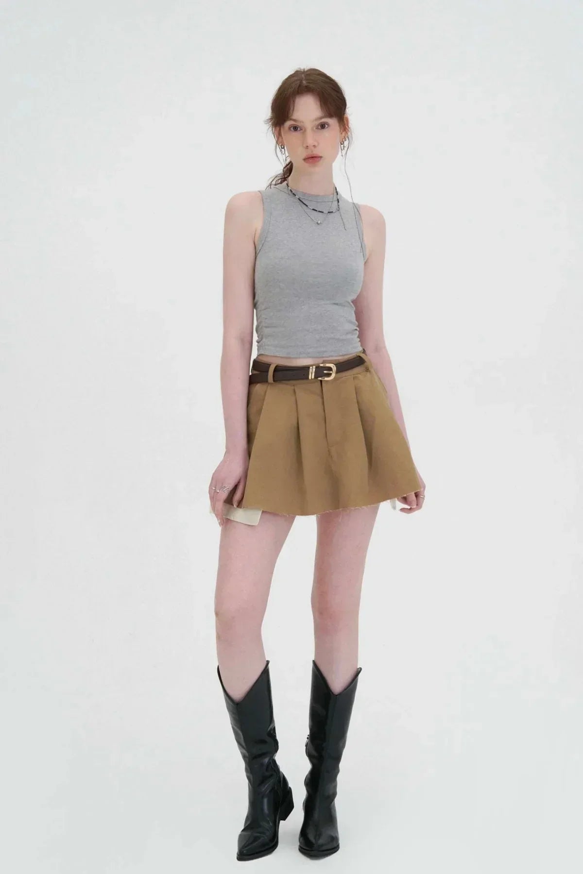 Woman wearing a beige wide tuck mini skirt with a black belt, gray sleeveless top, and black knee-high boots