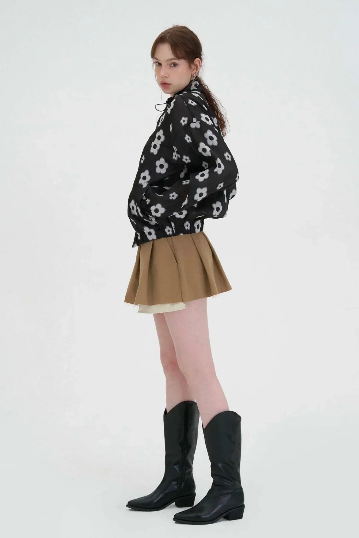 Side view of woman wearing black floral jacket, beige pleated mini skirt, and black knee-high boots