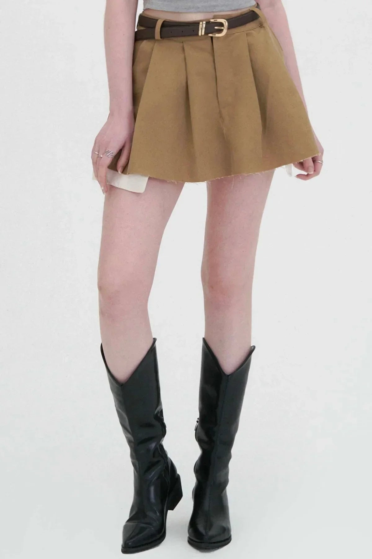 Woman wearing beige wide tuck mini skirt with brown belt and black knee-high boots on white background
