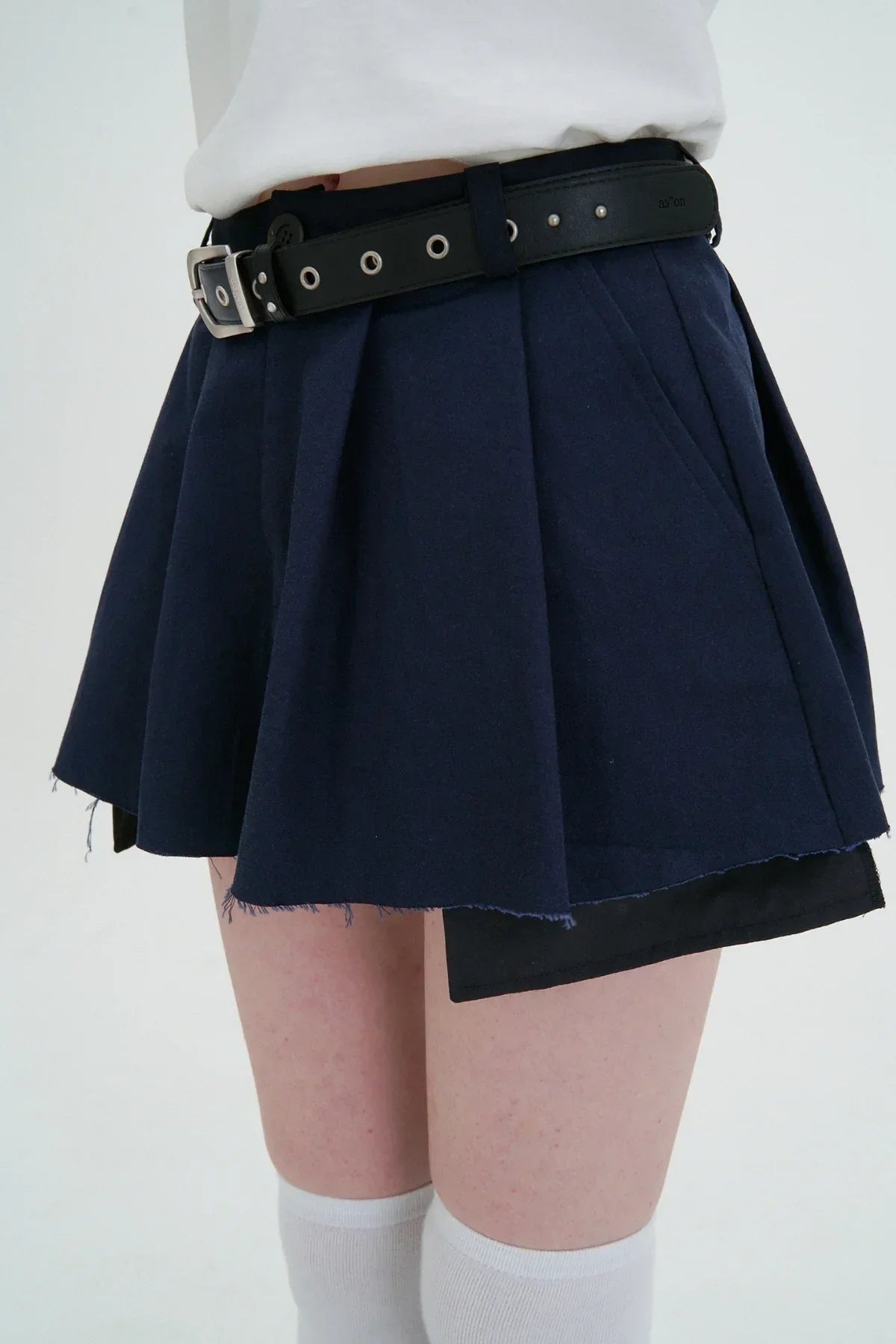 Close-up of a navy wide tuck mini skirt with raw hem and black belt worn with white knee-high socks