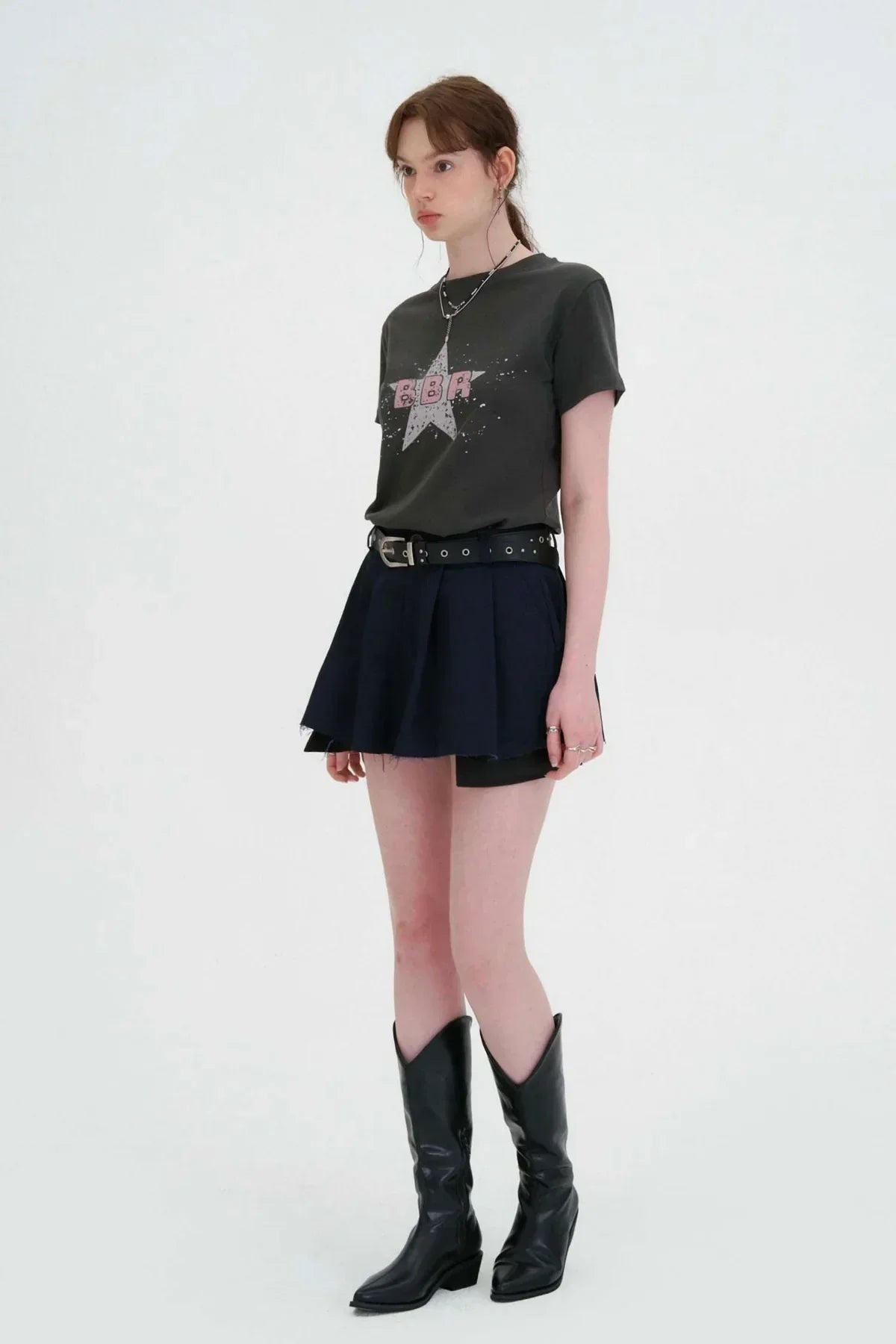 Model wearing black graphic tee, navy wide tuck mini skirt with black belt, and black cowboy boots